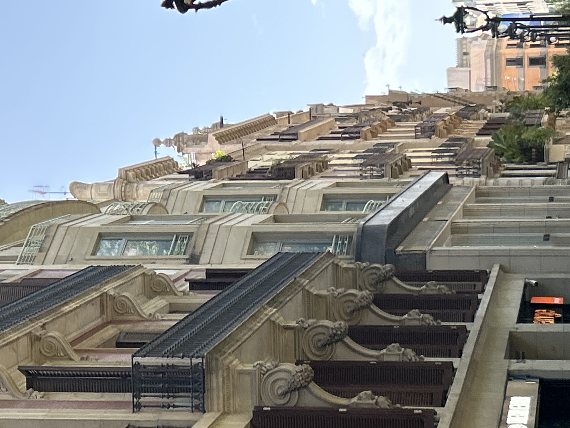 The image shows a view of a street in Barcelona, Spain, with traditional European architecture. On the left, a building with ornate balconies and shutters is visible. The balconies have dark metal railings and are supported by decorative stone brackets. The building's facade is a mix of cream and pink stripes. Further down the street, more buildings with similar architectural styles can be seen, featuring bay windows and multiple balconies. Some of the buildings have a pale yellow or light orange facade. The sky is clear and blue with a few white clouds. In the foreground, parts of the ground floor shops are visible, with one displaying a white sign with illuminated letters. A black lamppost with intricate detailing stands on the right side of the image. Lush green plants are visible on some of the balconies, adding a touch of nature to the urban landscape. The overall impression is of a sunny day in a historic European city.