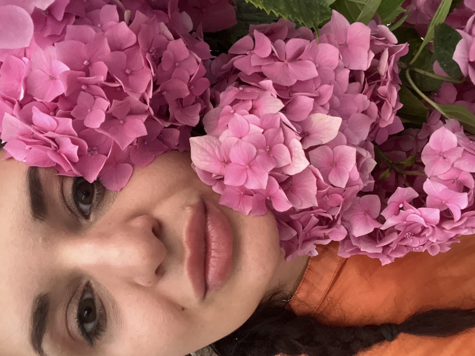 The image is a close-up, rotated photograph of a person's face adorned with pink hydrangeas. The subject's face is positioned horizontally, with their lips and part of their nose in the foreground, framed by the large, clustered pink blooms of the flowers. The hydrangeas are a vibrant pink, with multiple small petals forming each flower head. Some green leaves are visible amongst the flowers, suggesting they are fresh. The person appears to be looking upwards or away from the camera. Their skin is fair, and their lips are full and slightly parted, with a subtle sheen. Makeup is visible on their eyes, with eyeshadow and eyeliner. To the right of the person's face, a portion of an orange garment is visible, which appears to be a jacket or shirt. The background is out of focus, but hints of green from foliage and possibly other flowers are present. The lighting suggests it might be daytime, but the specific time of day and weather are not discernible. There is no visible text in the image. The overall impression is artistic and serene, with a focus on the interplay between the person's features and the natural beauty of the flowers.
