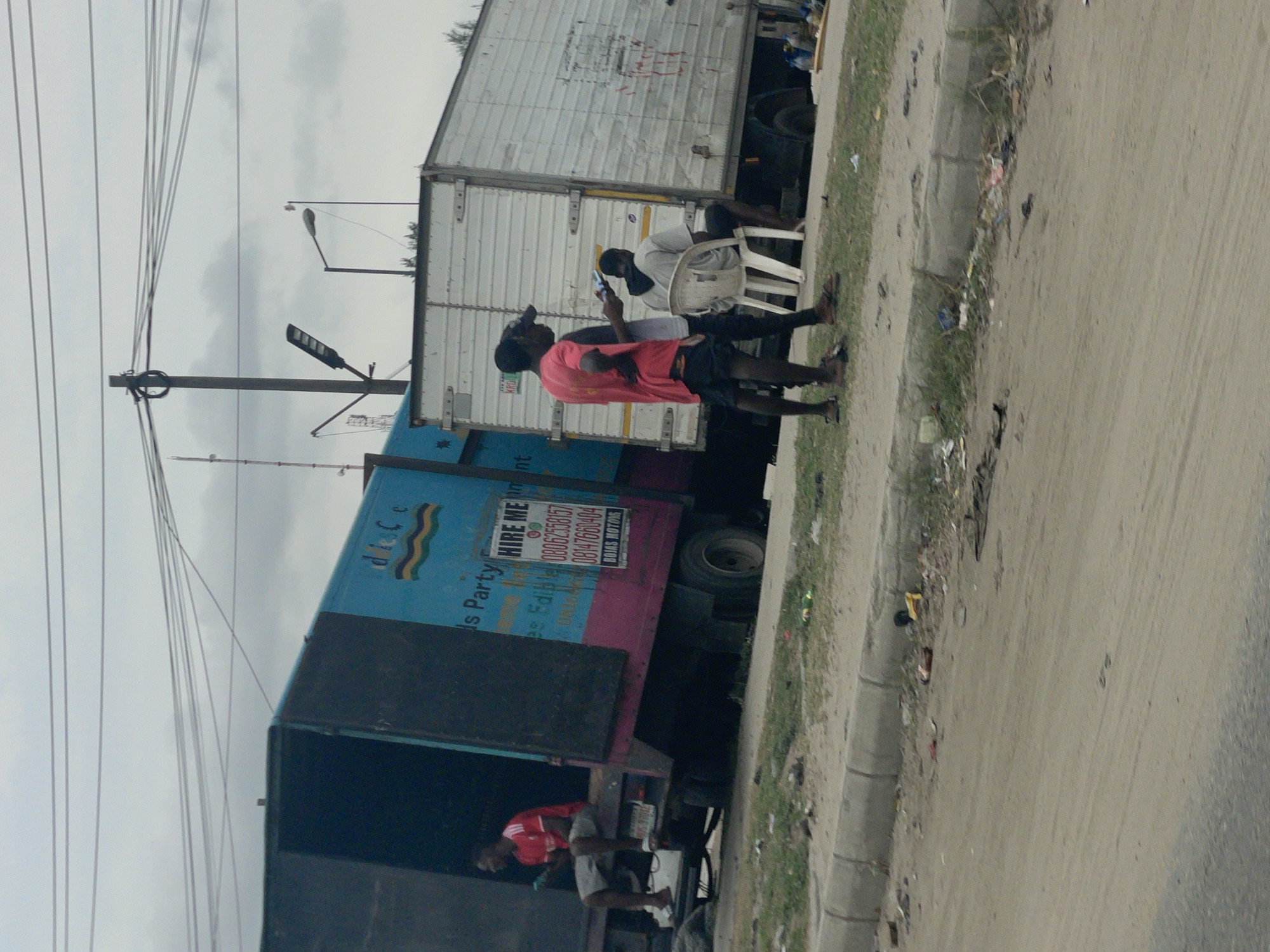 The image captures a street scene in Lagos, Nigeria, featuring two large trucks parked along a roadside. One truck has a white trailer, and the other is painted with blue and pink colors and has "Hire Me" in a sign. Three men are visible: one sitting on a plastic chair using a phone, another standing next to the seated man looking at a phone, and the third sitting inside the open back of the blue truck. Overhead, power lines stretch across the sky, and streetlights are also visible. The roadside is littered with debris.