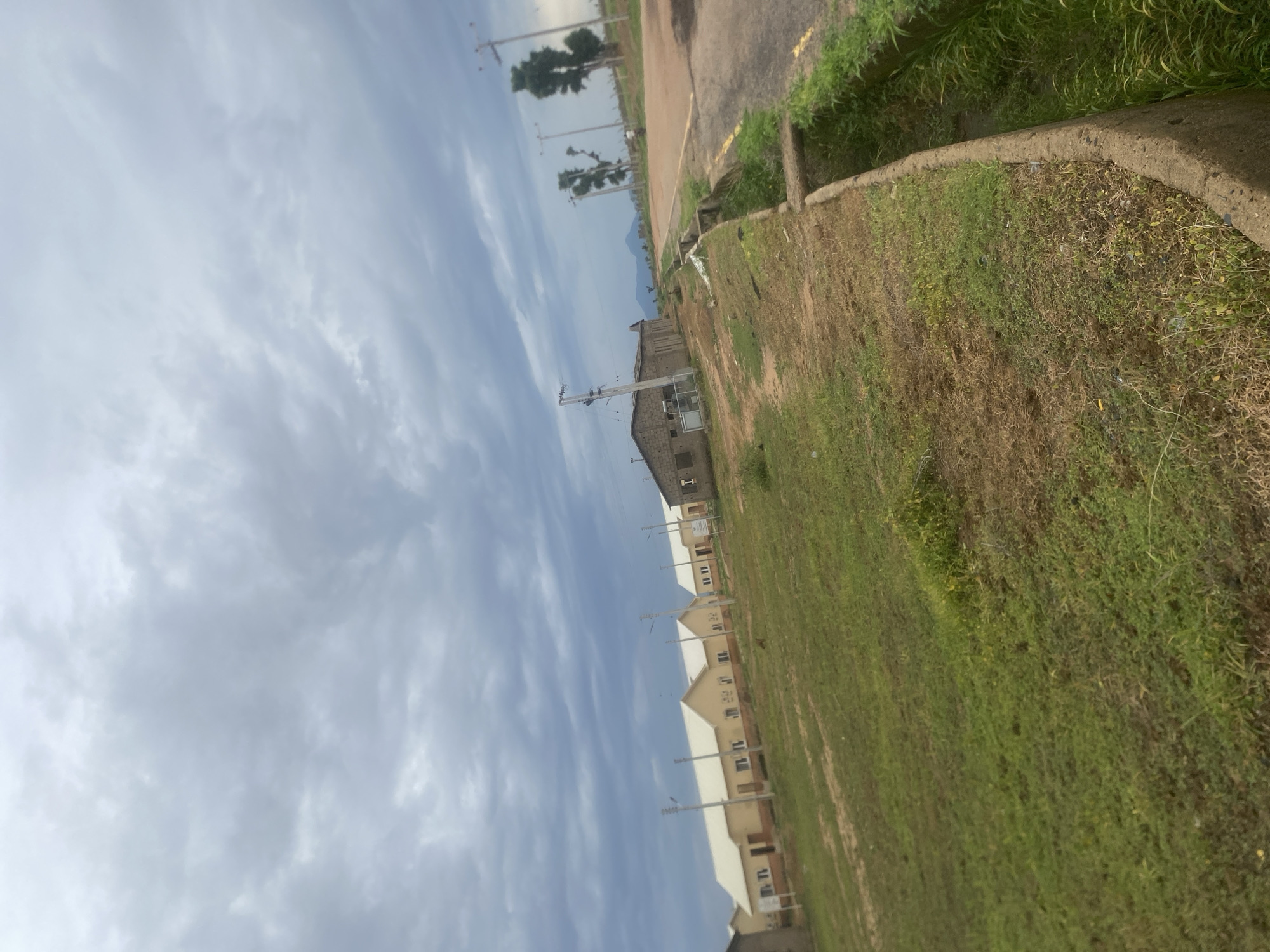 The image captures a partially constructed residential area in Bulamare, Nigeria. The scene is viewed from a low angle, looking upwards towards a cloudy sky. In the foreground, a grassy area with patches of dry earth slopes away, bordered by a concrete curb and a paved road with some overgrown vegetation along its edge.
Several newly built houses are visible in a line, their pale exteriors and white roofs contrasting with the natural surroundings. Some of the houses appear to be in the early stages of construction or finishing. Electrical poles and wires are scattered throughout the scene, indicating the presence of infrastructure. In the distance, silhouetted trees and what appears to be a mountain range can be seen against the sky, suggesting a rural or semi-rural setting. The lighting and cloudy sky suggest it is daytime, possibly late afternoon, with overcast weather. There is no discernible activity or people in the image. No text is visible.