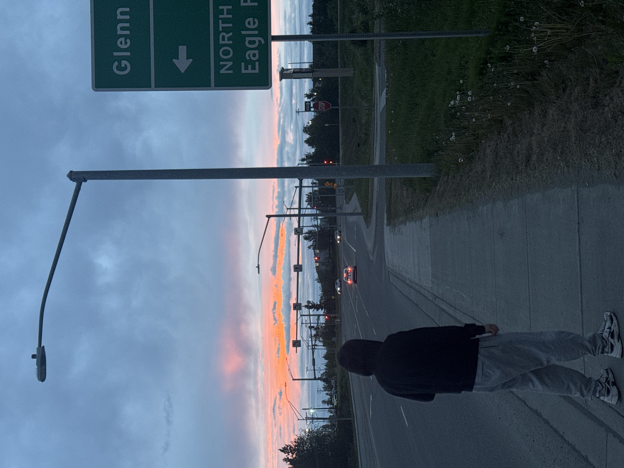 The image depicts a street scene at dusk in Anchorage, Alaska. A person wearing a black hooded sweatshirt and grey sweatpants stands on a sidewalk, facing away from the camera and towards a road. The sky is filled with colorful clouds, displaying hues of orange, pink, and blue, indicating either sunrise or sunset. A street light looms over the sidewalk. Across the road, traffic lights are visible, and several cars are driving. A green directional sign with white lettering is mounted on a pole, indicating "Glenn" with an arrow pointing left and "NORTH Eagle P" below. A stop sign is also visible further down the road. The environment is characterized by asphalt roads, sidewalks, grassy areas with dandelions, and trees.