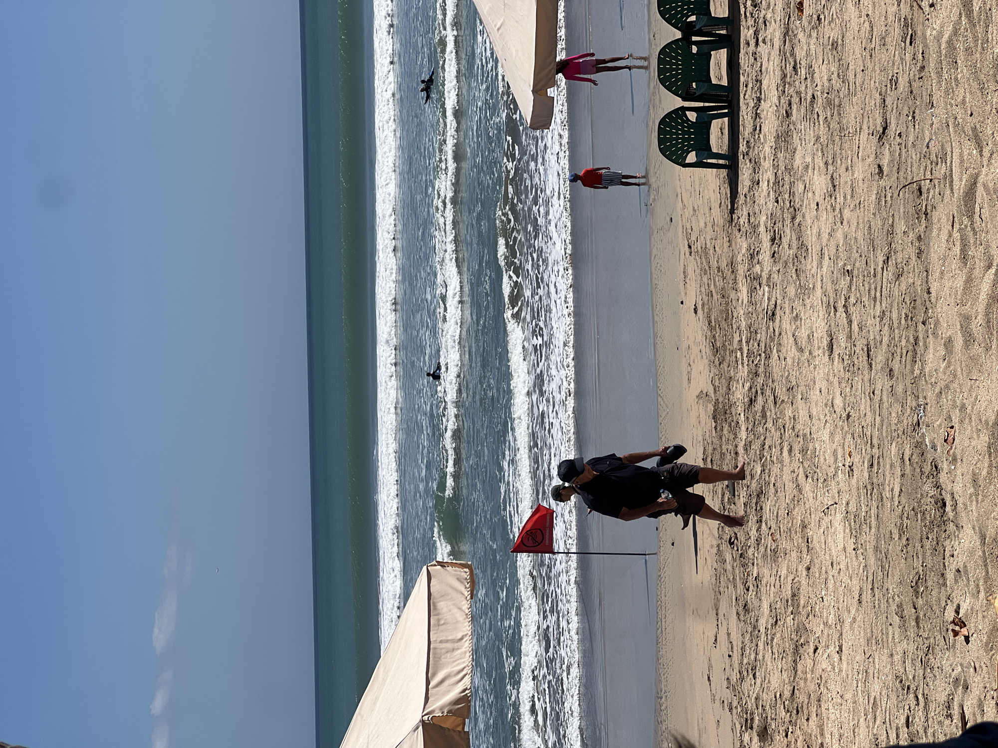 A bright, sunny day at Kuta Beach, Indonesia, with moderate waves rolling in. In the foreground, a sandy beach with several plastic green chairs is visible, along with a large beach umbrella on the left. A man in a black shirt and shorts walks away from the camera, carrying a bottle and a hat. Near him, a red flag with a symbol of a person being pulled by waves is planted in the sand, indicating dangerous swimming conditions.
Further down the beach, a young boy wearing a red shirt and striped shorts stands facing the ocean. To his right, a young girl in a pink top and shorts is also looking at the sea. In the middle distance, several surfers are in the water, riding or waiting for waves. A second beach umbrella is visible on the right side of the image. The sky is clear blue with a few wispy clouds. The overall impression is a lively beach scene with both leisure activities and safety warnings.