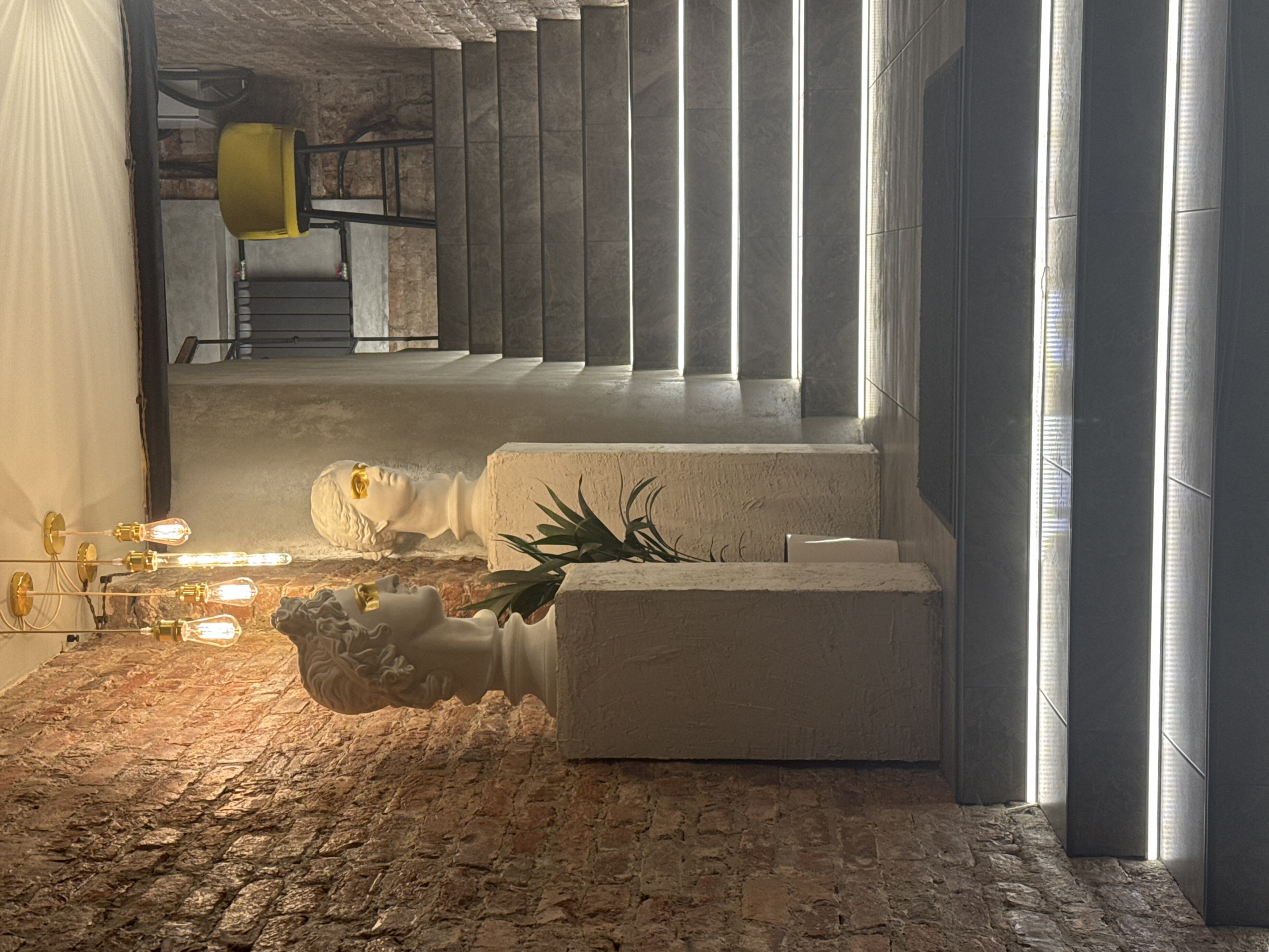 The image captures an interior space featuring a brick floor, two classical-style busts placed on pedestals, stairs, and vertical lighting elements. The busts, depicted in white with gold accents, are positioned on two rectangular pedestals which also hold green plants. A chandelier is fixed to the wall in the left corner. Above the busts and pedestals, there are steps leading upwards, and beside the steps, illuminated vertical lighting panels run from the floor to the ceiling. A yellow chair and other furniture are partially visible in the background near the stairs. The scene is located in the city of Saint Petersburg, Russia.