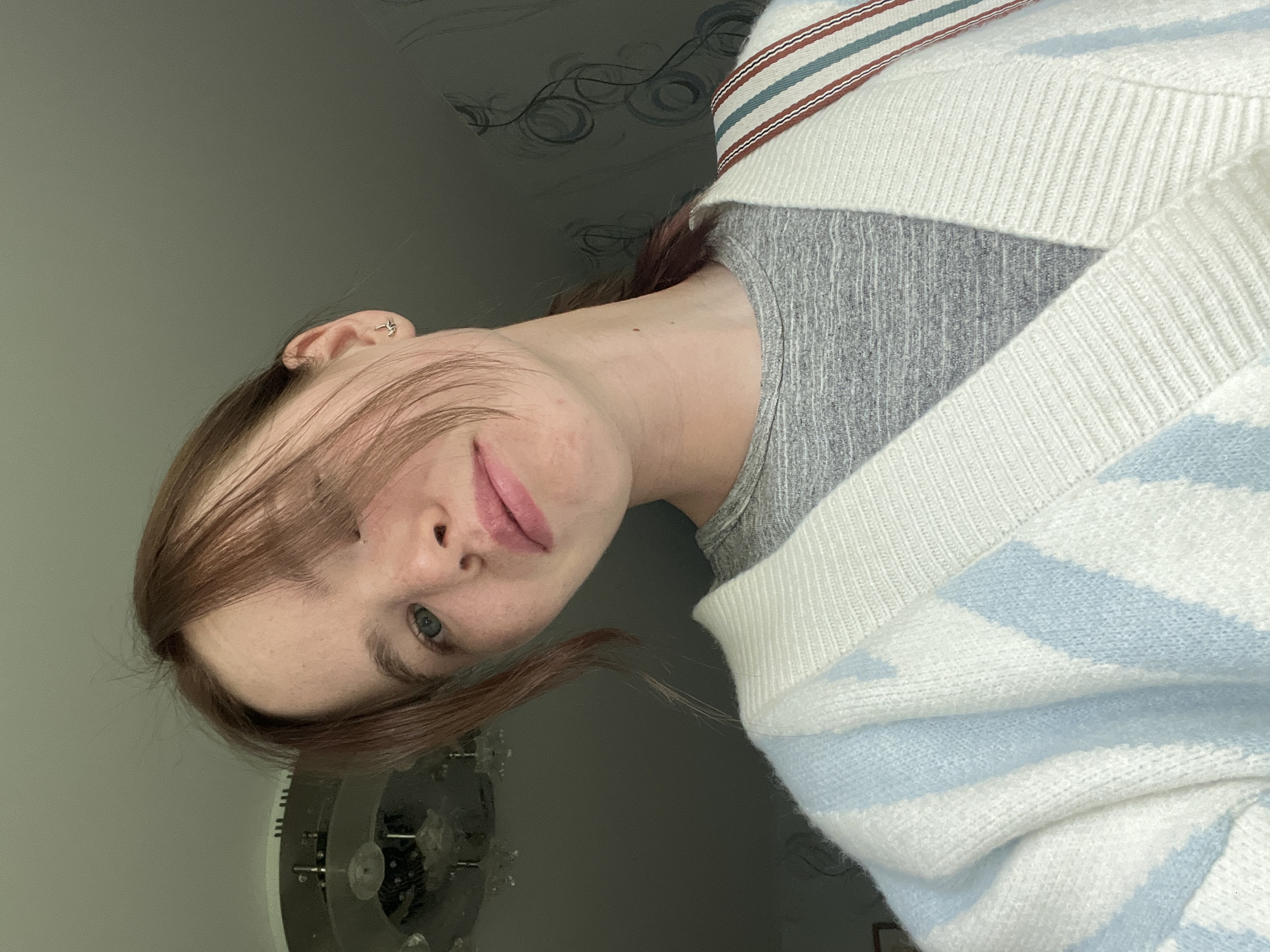 A young woman with reddish-brown hair, blue eyes, and fair skin is the primary subject of the photograph. She is wearing a gray top and a white and light blue striped sweater. Her hair partially covers her face. She is looking directly at the camera. The background includes a wall with a patterned design and a decorative object that appears to be a fan or lighting fixture. The setting is in an interior, presumably in the city of Chaikovsky, Russia.