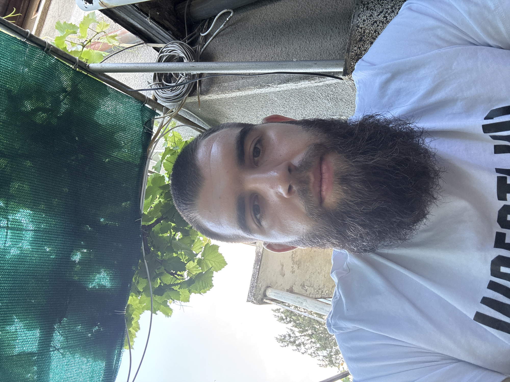 A medium shot features a man with a thick, dark beard and short hair. He is wearing a white t-shirt with a large, dark text on the front. The man is looking into the camera. The setting appears to be outdoors, possibly in a backyard or patio. There's a green mesh shade structure overhead with vine leaves visible. Behind the man, the background shows a textured concrete wall and some cabling. Based on visual cues and the prompt's direction, the location is Novoselets, Bulgaria.