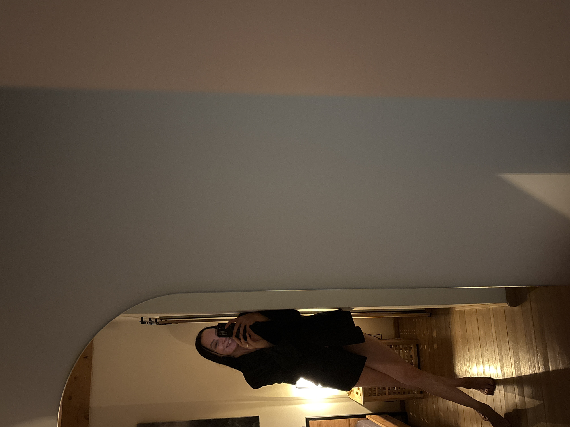 A woman is visible in a mirror. She is holding a phone in front of her, presumably taking a picture of herself. She appears to be wearing a black robe or dress. The reflection shows a room with wooden floors. The location is unknown and the image does not provide any evidence suggesting this location is Chaikovsky, Russia.