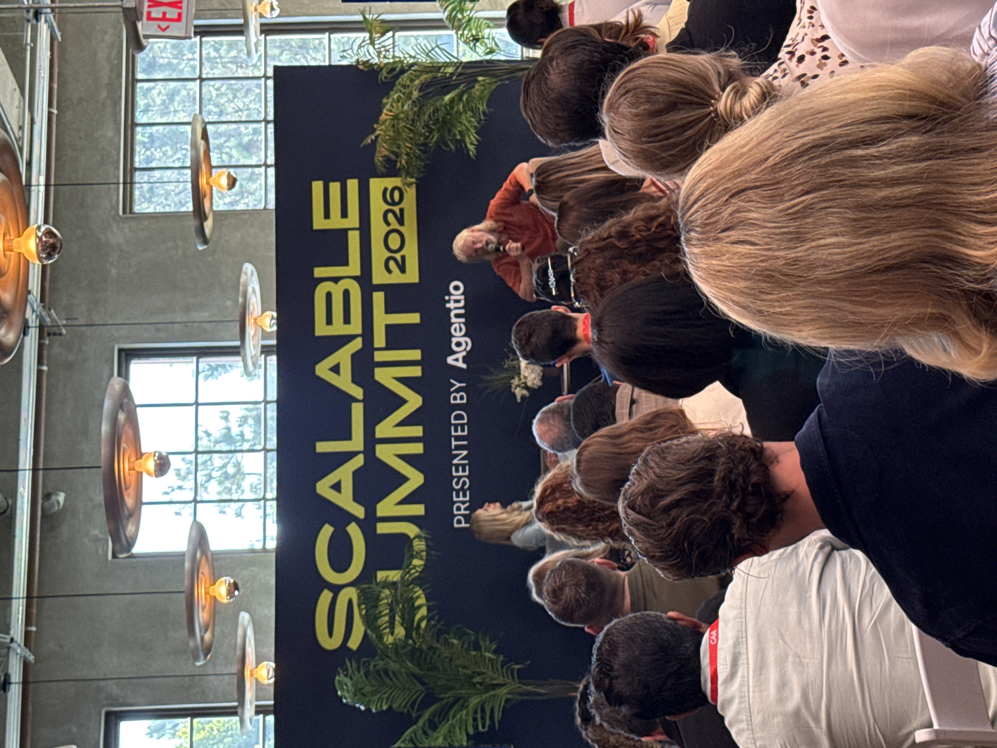 The image depicts an indoor conference setting with a group of people seated and facing a speaker on a stage. A banner behind the speaker reads "Scalable Summit 2026 Presented by Agentio". The room has a modern, industrial-style aesthetic with large windows, exposed piping, and unique pendant lighting. The attendees are of various ages and genders, and they are focused on the presentation. Some are wearing badges or lanyards. The location is implied to be in Los Angeles, United States of America, based on the summit year displayed in the background.