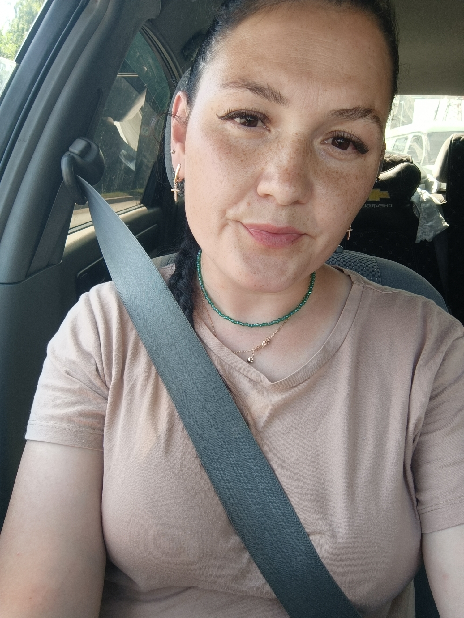 A woman is seen from the chest up, inside a car. She is wearing a light brown t-shirt and a grey seatbelt is diagonally across her body. She has dark hair pulled back in a braid and wears a green beaded necklace with a small silver heart charm. She has freckles across her nose and cheeks. The background is out of focus, showing the interior of the car, including a car seat headrest with the word "CHEVROLET" visible. Sunlight is visible through the car window, suggesting it is daytime. The location of Chaikovsky, Russia, is not ascertainable from the image.