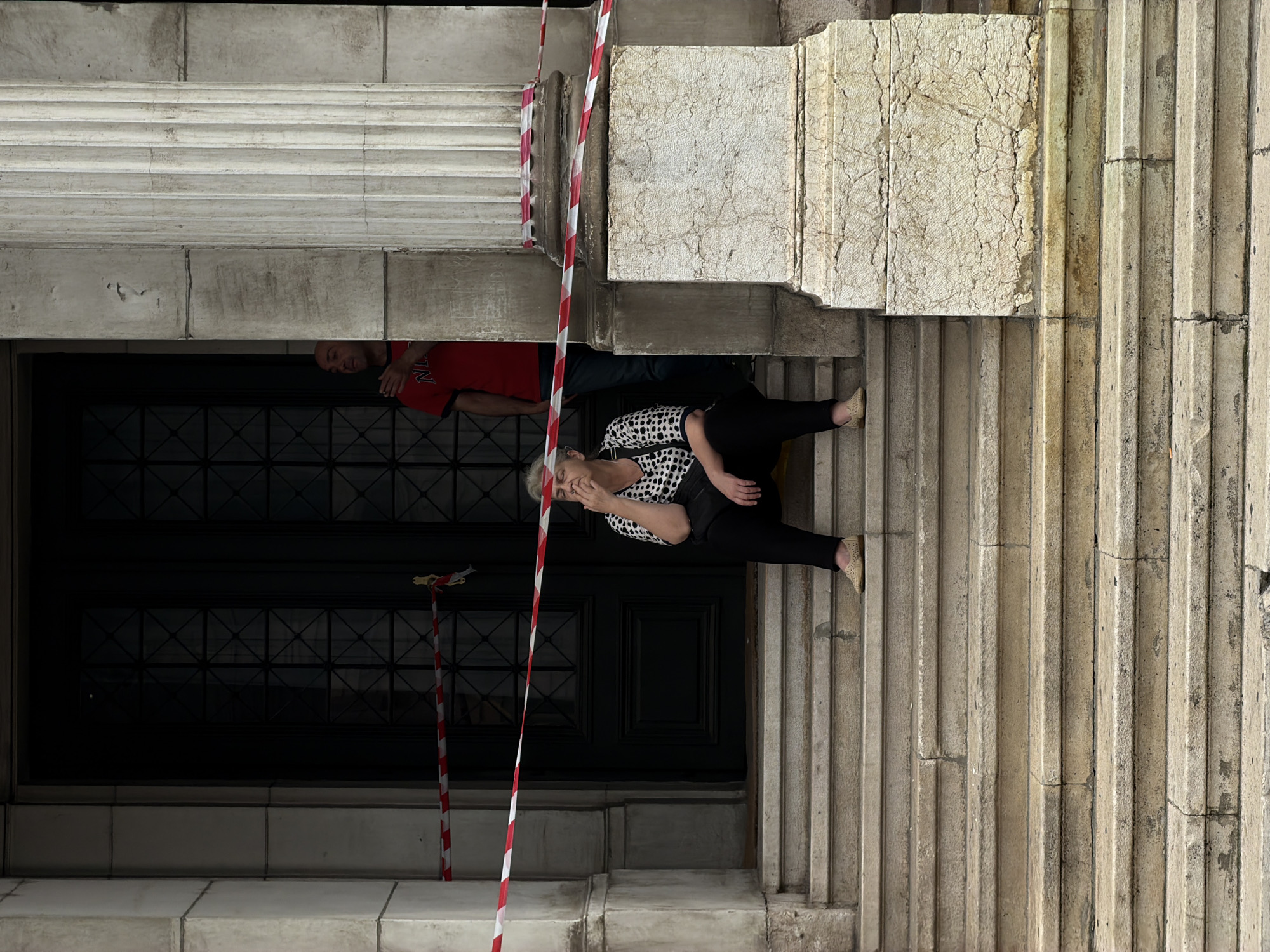 The image depicts an outdoor scene on a stone staircase in front of a building. Red and white caution tape is strung across the entrance of the building and over the first few steps of the staircase. A woman with grey hair is seated on the steps, wearing a black and white patterned top and black pants. She is holding what appears to be a cigarette to her mouth. Behind her, a man wearing a red t-shirt is visible standing within the doorway of the building, partially obscured by a large stone pillar. The building has a grand entrance with large dark double doors. The architectural style suggests it could be a historical or institutional building. The presence of caution tape implies the area may be cordoned off for some reason, such as ongoing construction, maintenance, or a security incident. The overall atmosphere is somewhat somber, with the woman's posture and the caution tape contributing to this impression. The location is identified as Sarajevo, Bosnia and Herzegovina.