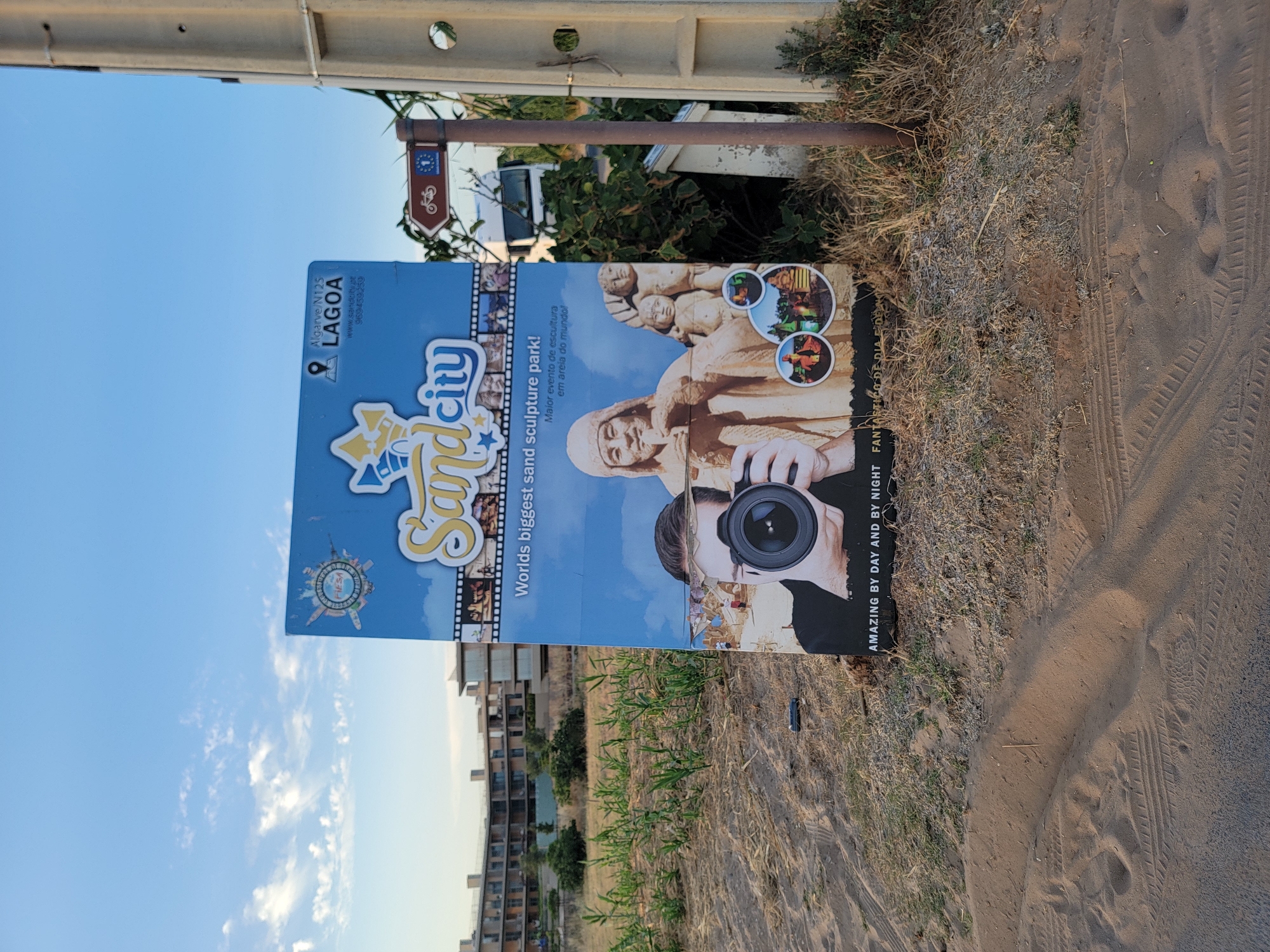 The image shows a large advertisement for "Sand City," a sand sculpture park in Lagoa, Portugal, located on the Algarve coast. The advertisement features vibrant graphics and text, promoting the park as the world's biggest sand sculpture park. In the foreground, a man is depicted holding a camera, with a sand sculpture of a child and other figures visible in the background. The advertisement is placed on a sandy, dry terrain, with sparse vegetation and a building complex in the distance, suggesting a coastal or resort area. A directional signpost to the right of the advertisement indicates a bicycle route and the European Union flag. The sky above is clear blue with scattered white clouds, indicating fair weather. The overall scene captures a promotional display for a tourist attraction in a natural outdoor setting.