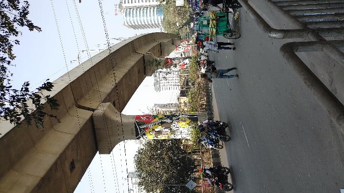 The image depicts a daytime street scene in the Kawran Bazar area of Dhaka, Bangladesh, beneath the elevated viaduct of the Dhaka Metro Rail (MRT Line 6). A large, light-colored concrete metro rail viaduct spans the upper and central portions of the frame, supported by prominent pillars adorned with colorful, abstract murals. Numerous overhead utility wires crisscross the sky.
The asphalt road, visible in the mid-ground, shows multiple lanes of traffic. Several vehicles and pedestrians are present. On the left side of the road, at least five motorcycles are in motion; one red motorcycle carries two riders, the front rider wearing a green helmet and backpack. A blue motorcycle and a dark-colored motorcycle are also visible with single riders. In the mid-ground on the right, a male pedestrian in a light-colored shirt and cap holds a mobile phone to his ear while walking. Another male pedestrian in a plaid shirt walks beside him. A green, two-wheeled handcart, laden with green produce (likely bananas or plantains), is positioned on the far right side of the road, operated by an individual.
In the background, multiple multi-story buildings rise, including a distinctive modern high-rise with a curved facade labeled "BONAE" (Borak Tower) on the far right. Tree branches with dark green leaves partially obscure the viaduct in the upper left, and sparse vegetation lines the road median. A metal guardrail extends across the lower right foreground. The sky is bright, appearing clear to hazy.