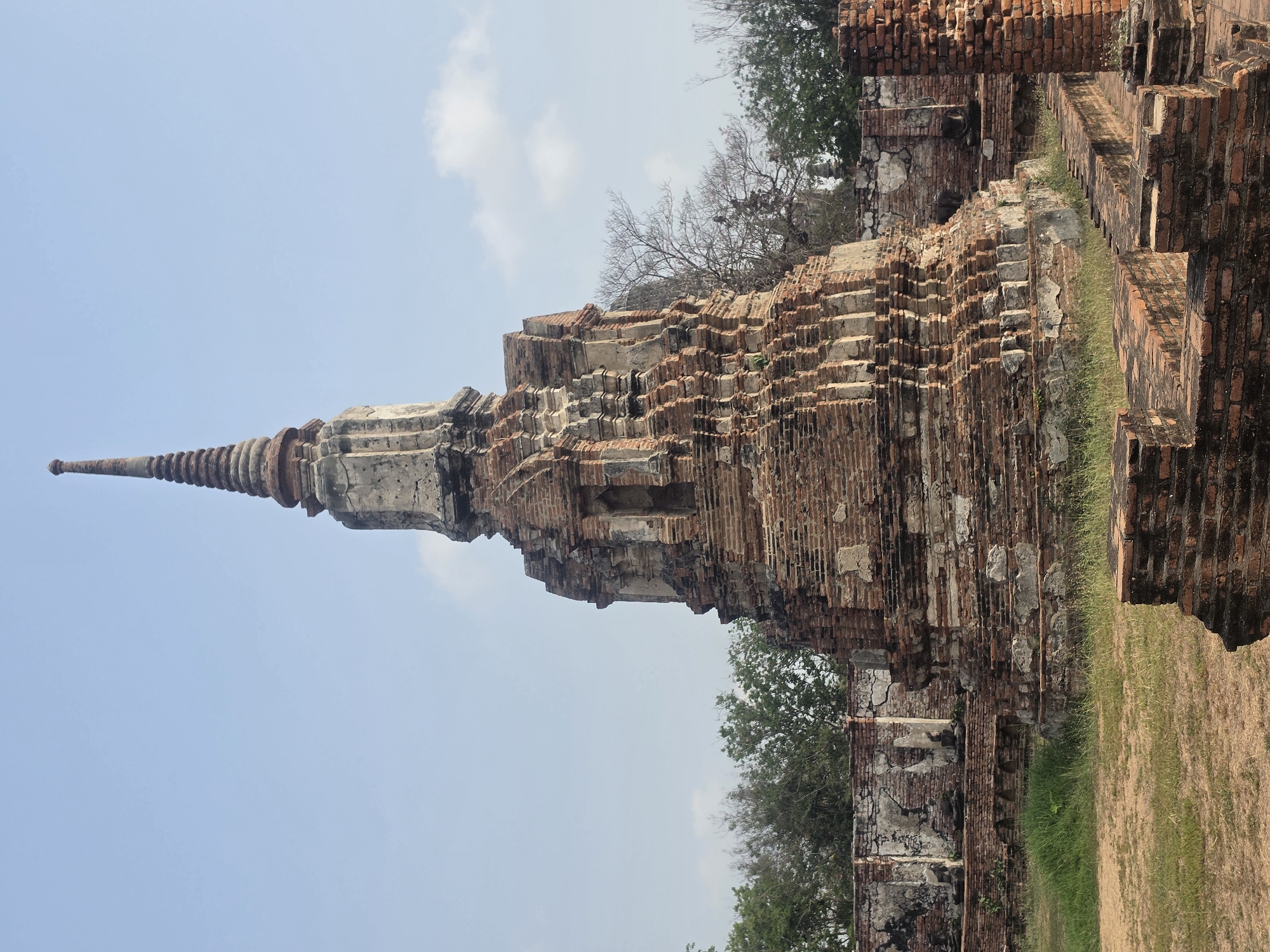 The image captures a towering, ancient brick structure, likely a stupa or chedi, amidst the historical landscape of Phra Nakhon Si Ayutthaya City Municipality, Thailand. The structure is predominantly constructed of weathered, red-brown bricks, exhibiting a tiered design that culminates in a pointed spire. Evidence of age and decay is visible throughout the structure, with missing bricks and exposed inner layers. Patches of greenery, including grass and small trees, surround the base of the structure. The sky is a pale blue, with scattered clouds providing a muted backdrop.
