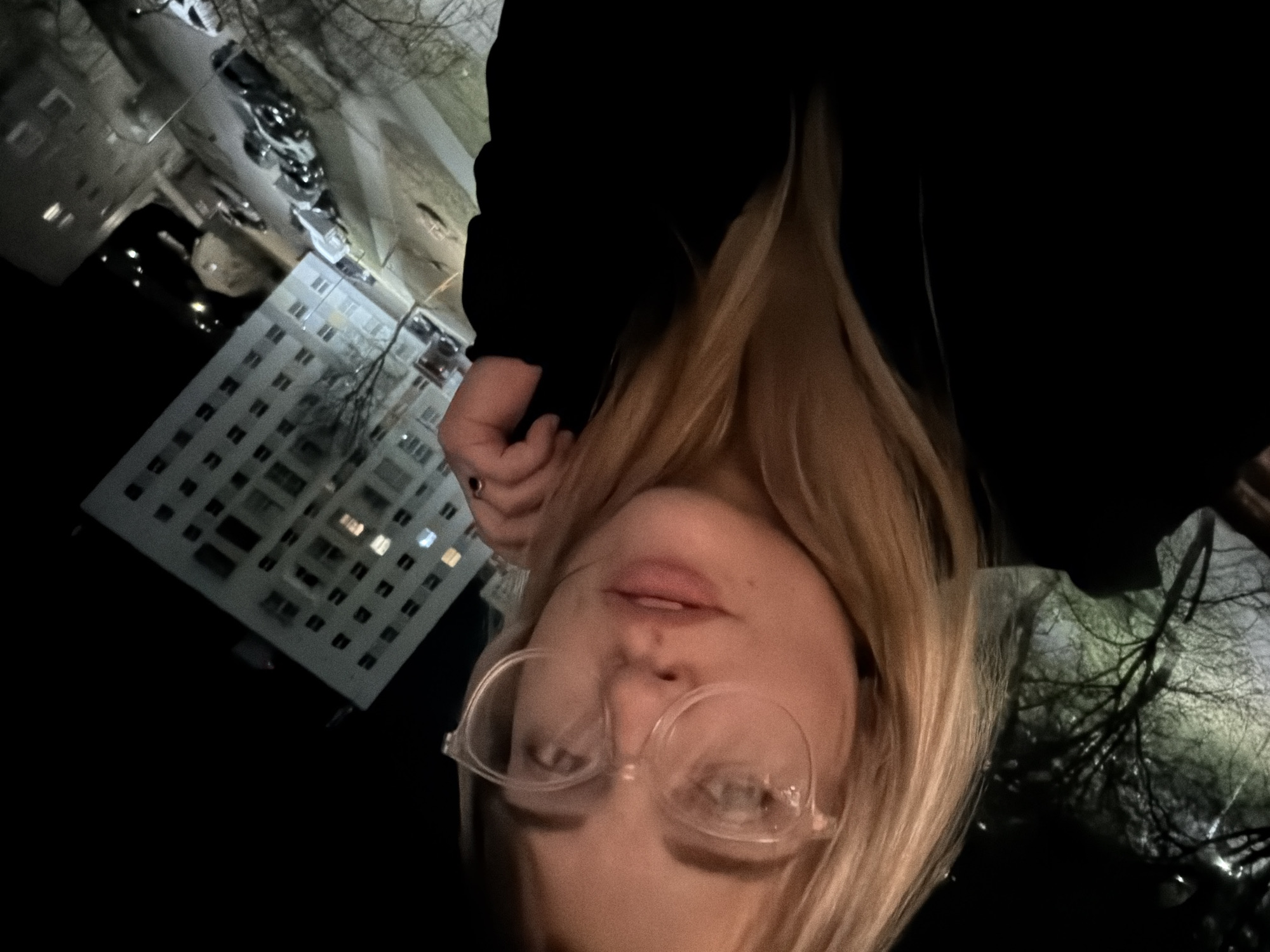 A nighttime selfie depicts a young, fair-skinned person with blonde hair and glasses. The subject is wearing a black garment. The background reveals an urban landscape featuring a multi-story building with illuminated windows. Above the building is a street with parked cars. Bare trees are visible, with light reflected on their branches. The photograph seems to be taken from an elevated position, facing downward. Based on building architecture and car models, the location is most likely the city of Naberezhnye Chelny, Russia.