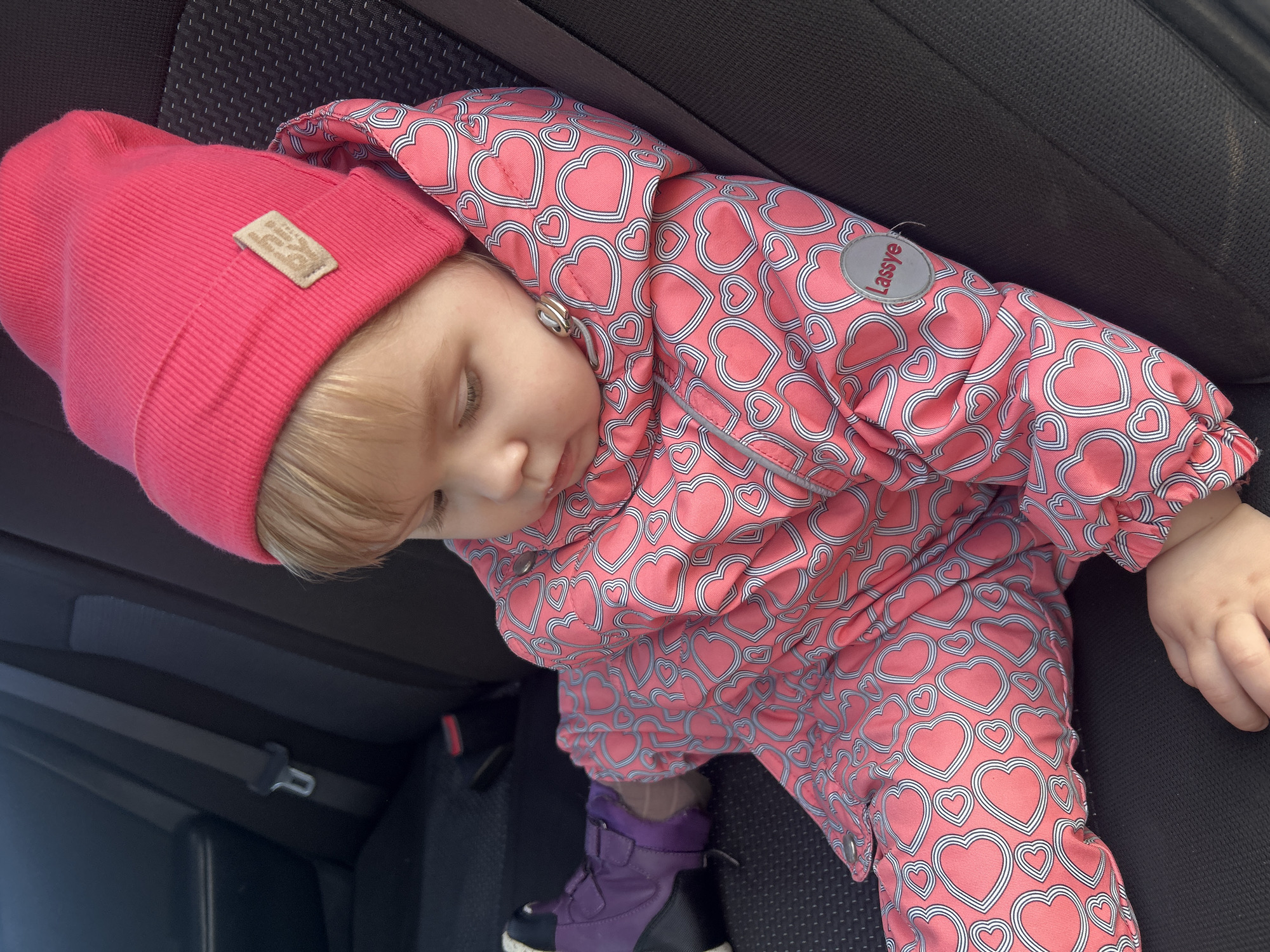 A young child wearing a pink beanie and a pink heart-patterned snowsuit is asleep in a car seat. The child is wearing purple boots and is buckled into the black car seat. The brand "Lassie" is visible on the snowsuit. The location is in a vehicle, but the city of Ольховка, Russia, is not confirmed from the image.
