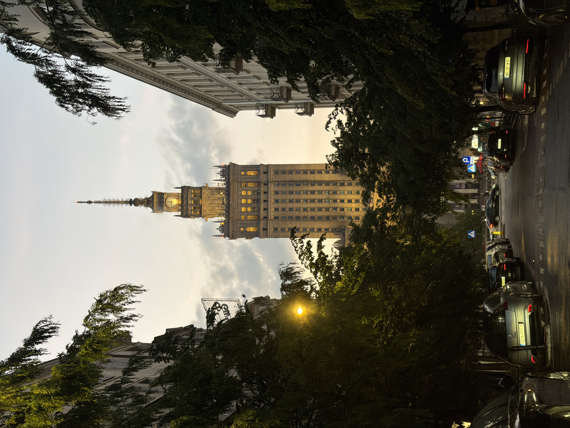 The image is an eye-level outdoor shot of the Palace of Culture and Science in Warsaw, Poland, framed by trees and buildings on either side. The street, lined with parked cars, recedes into the distance towards the palace. The sky is overcast with an early evening glow behind the building. The palace is illuminated with lights on.