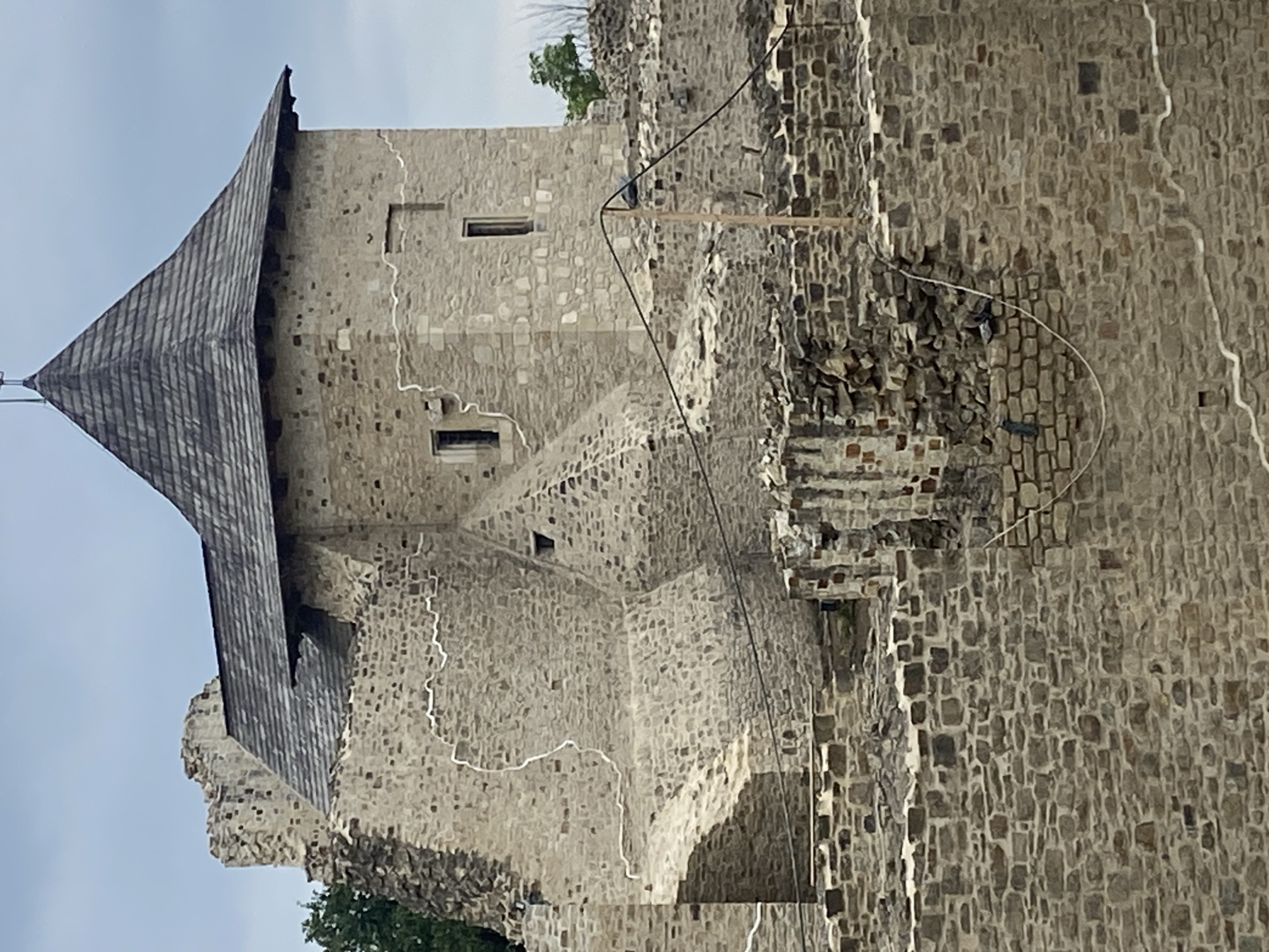 The image shows a partial view of the ruins of the Stephen the Great's Fortress in Suceava, Romania. The focus is on a stone tower with a wooden shingled roof and a smaller adjacent structure, also made of stone and partially ruined. The walls are constructed from irregular stone blocks of varying sizes and colors, with some evidence of plaster or repair work visible as white lines on the surfaces. Several small, dark rectangular openings, likely windows or arrow slits, are present on the tower. The surrounding area is characterized by more stone ruins, suggesting a larger complex that has undergone significant decay. A wooden post with wires running across it is visible in the foreground, partially obscuring some of the stone structures. The sky is overcast with a pale blue and gray hue. No people are visible in the frame.