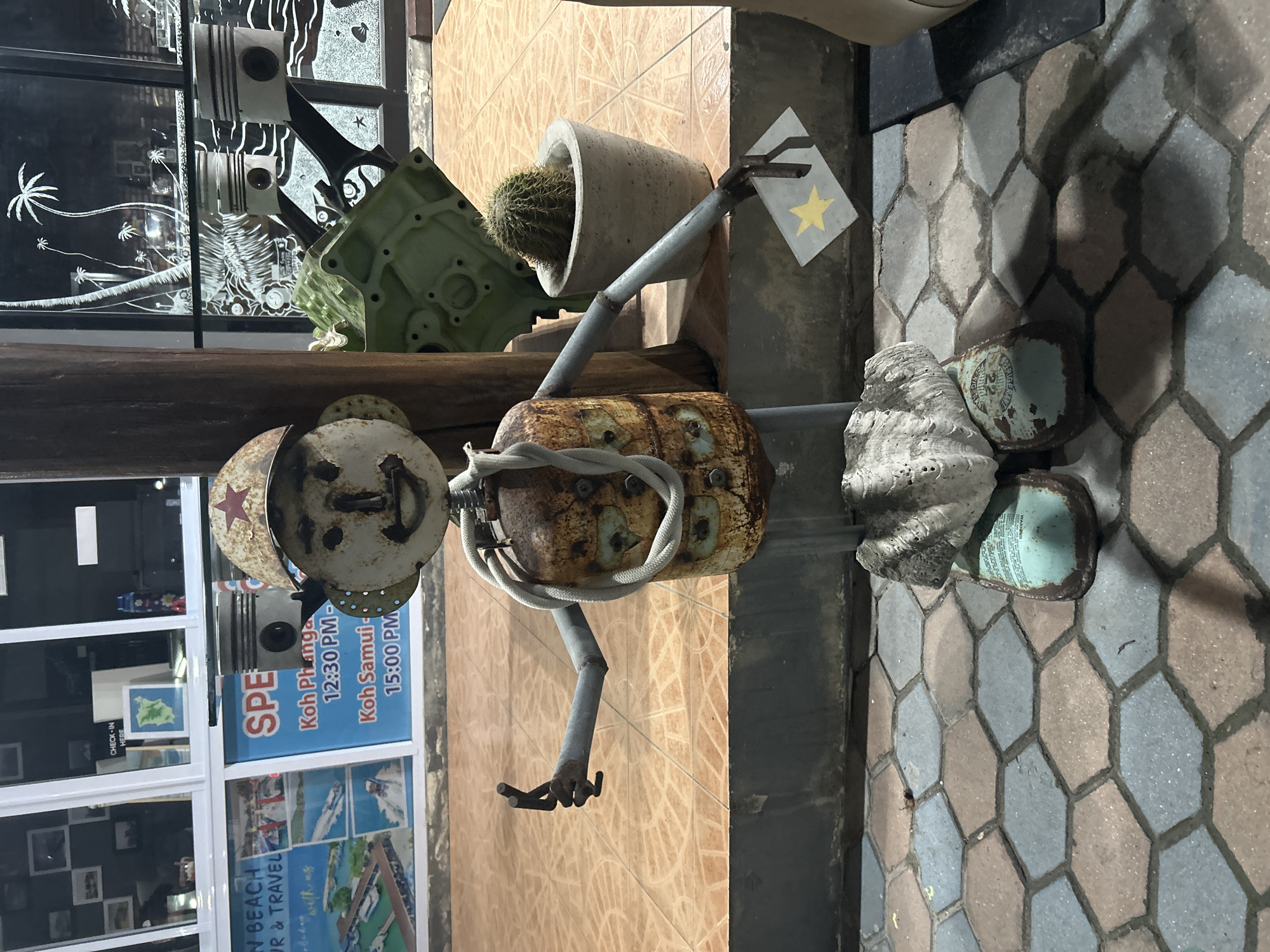 The image captures a whimsical outdoor scene in Ban Haad Rin, Thailand. It features a handmade metal sculpture of a person, constructed from various repurposed materials. This figure has a round head with a rusty finish, topped with a metal hat adorned with a red star. Its body is a cylindrical metal container, bound with a white rope. The arms appear to be made from metal rods, one of which is holding a small, square piece of metal painted with a yellow star. The figure stands near a tiled wall, with a cactus in a concrete pot positioned above it.
Beside the sculpture are natural elements, including a large seashell and what appears to be a painted, used can. The ground is paved with hexagon-shaped tiles in varying shades of brown and grey.
In the background, a window displays advertising for travel between Koh Phanga and Koh Samui, with times listed as 12:30 PM and 15:00 PM, respectively. The travel agency name includes "BEACH TRAVEL." Reflections in the window reveal more of the surrounding environment and the artistry using car parts above.
