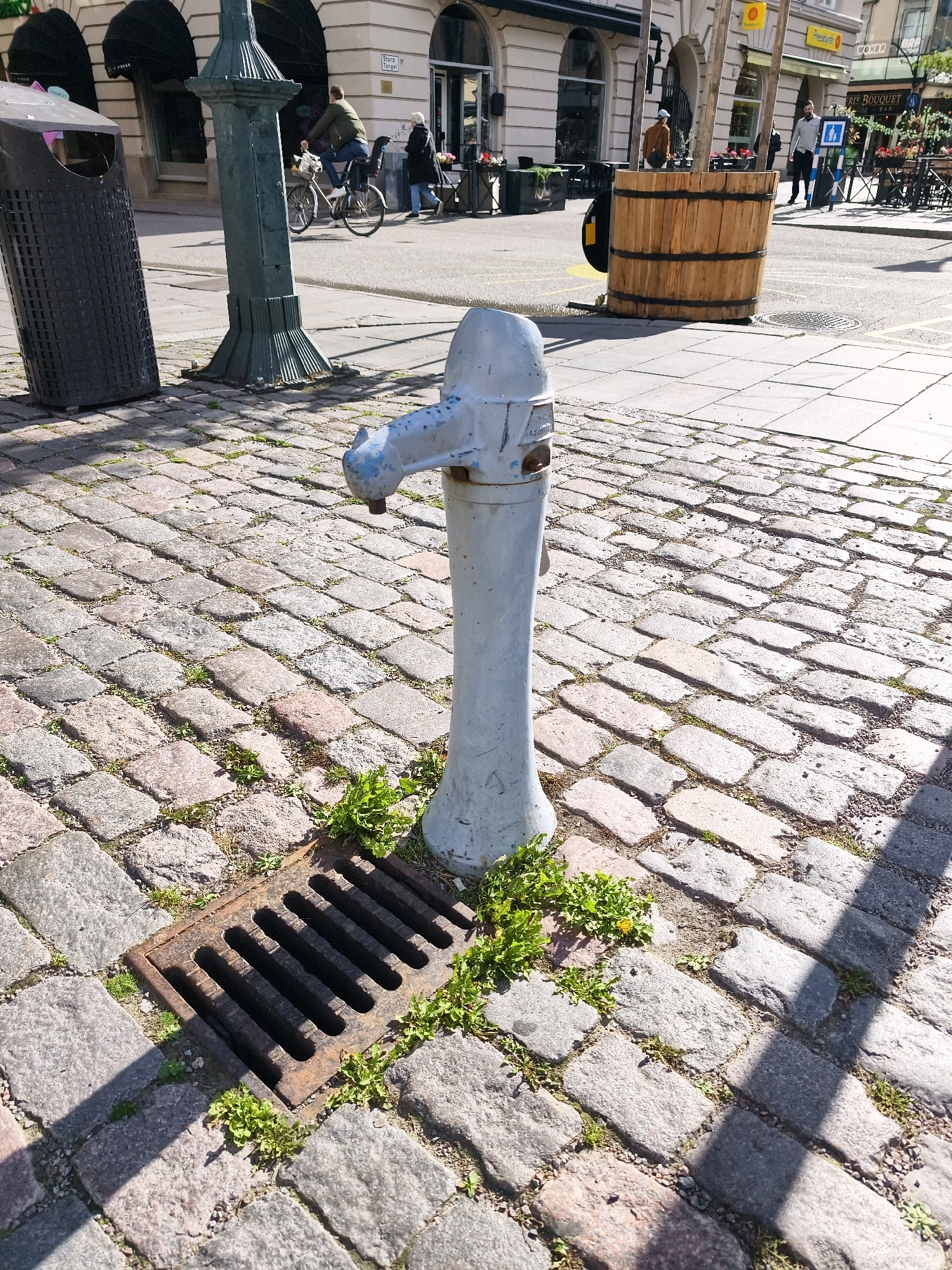 A light blue, antique water pump stands in the foreground on a cobblestone street. The pump has a rustic, aged appearance with some chipped paint. To the left of the pump, a dark grey trash receptacle is visible. In the mid-ground, a large, wooden barrel is situated on the street. Beyond the barrel, several people are seen walking and cycling. The background reveals buildings with awnings and outdoor seating areas, indicative of a commercial district. This scene is located on Stora Torget in Linköping, Sweden, as indicated by a sign on one of the buildings. The ground is paved with cobblestones, and some patches of green vegetation are growing through the cracks. The lighting suggests it is daytime with sunlight casting shadows.