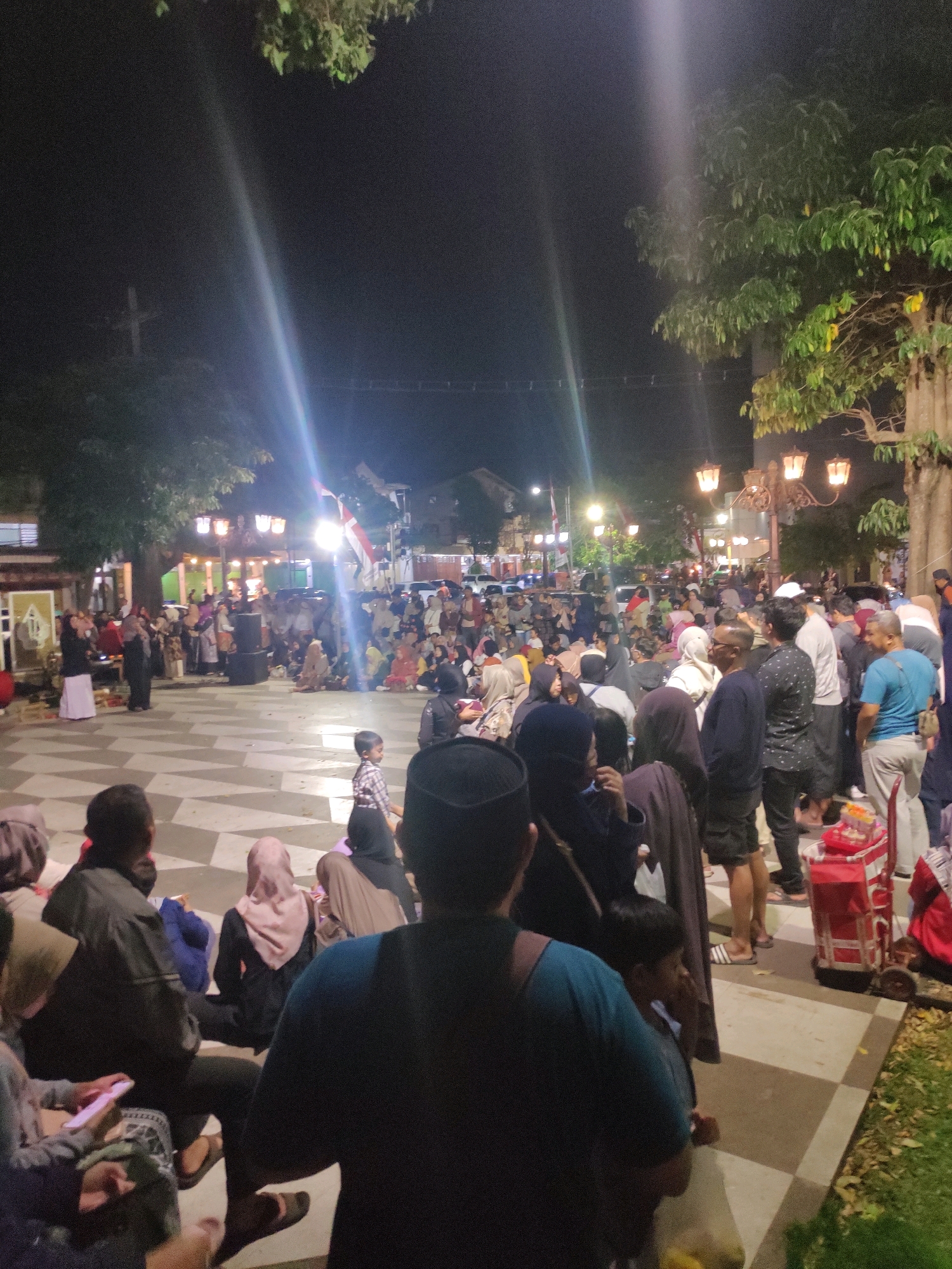 The image depicts a nighttime scene in a public square in Magetan, Indonesia. A large crowd of people, predominantly dressed in modest clothing and many wearing headscarves, are gathered in the square. The square's paving stones have a distinctive black and white checkered pattern. In the background, there are trees with illuminated lanterns hanging from them, casting a warm glow. A stage-like structure with speakers is set up, suggesting a performance or event is taking place. Several individuals are seated on the ground, while others stand and converse. A red food cart is visible on the right side of the image. The overall atmosphere appears to be lively and communal, likely a cultural or religious gathering.