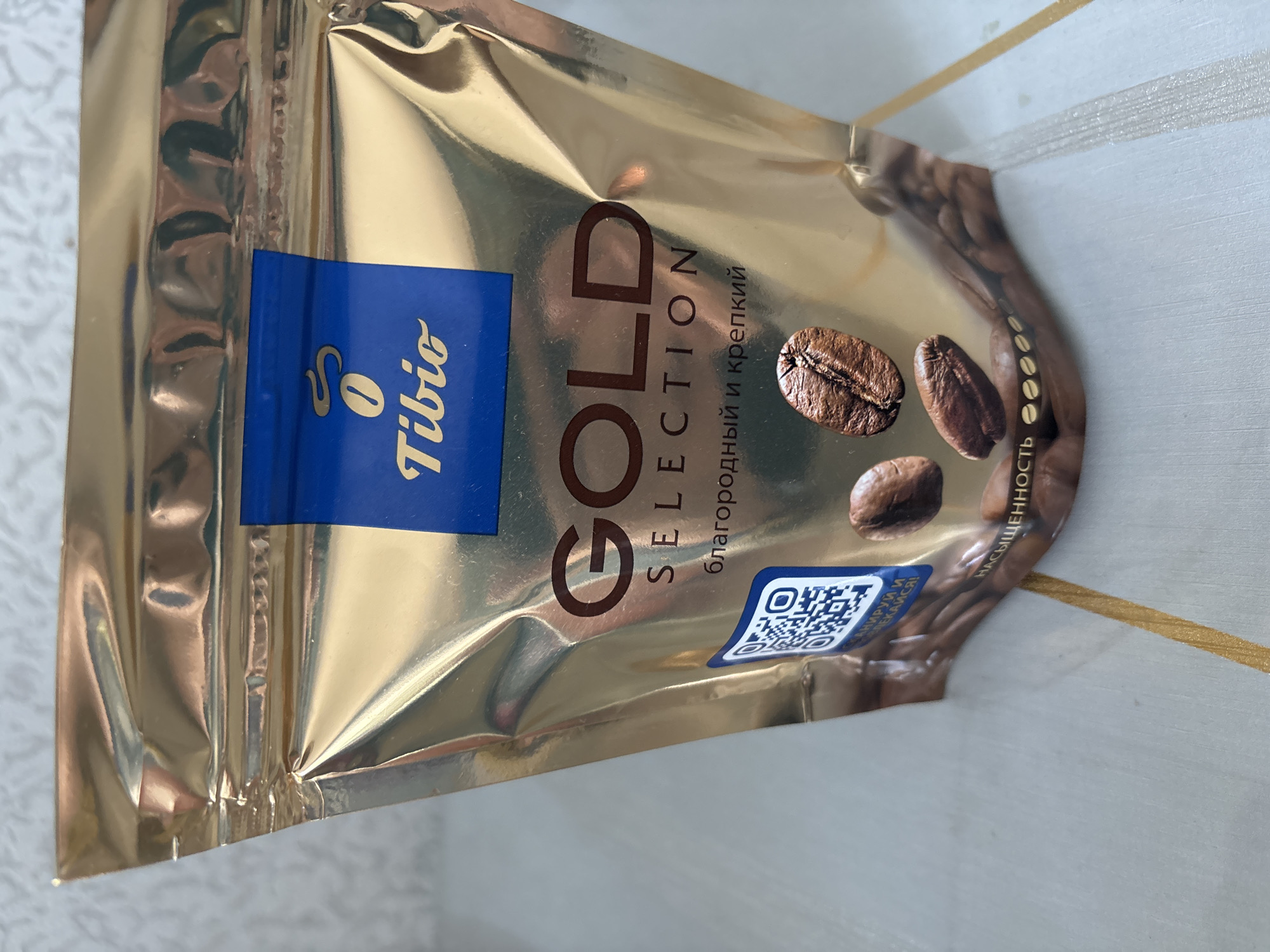 The image shows a close-up of a gold-colored foil package of Tibio brand coffee. The package is sealed at the top. The front of the package features the Tibio logo, which includes a stylized coffee bean, and the text "GOLD SELECTION" in large, dark brown letters. Below this, there is text in Russian that translates to "noble and strong." A QR code is visible on the left side of the package. Several coffee beans are depicted on the packaging. The package is placed on a surface that appears to be a light-colored table with a striped pattern. The background is out of focus and appears to be a light-colored textured surface. There are no people visible in the image. The scene is not identifiable as being in Chernogorsk, Russia or any other specific city.