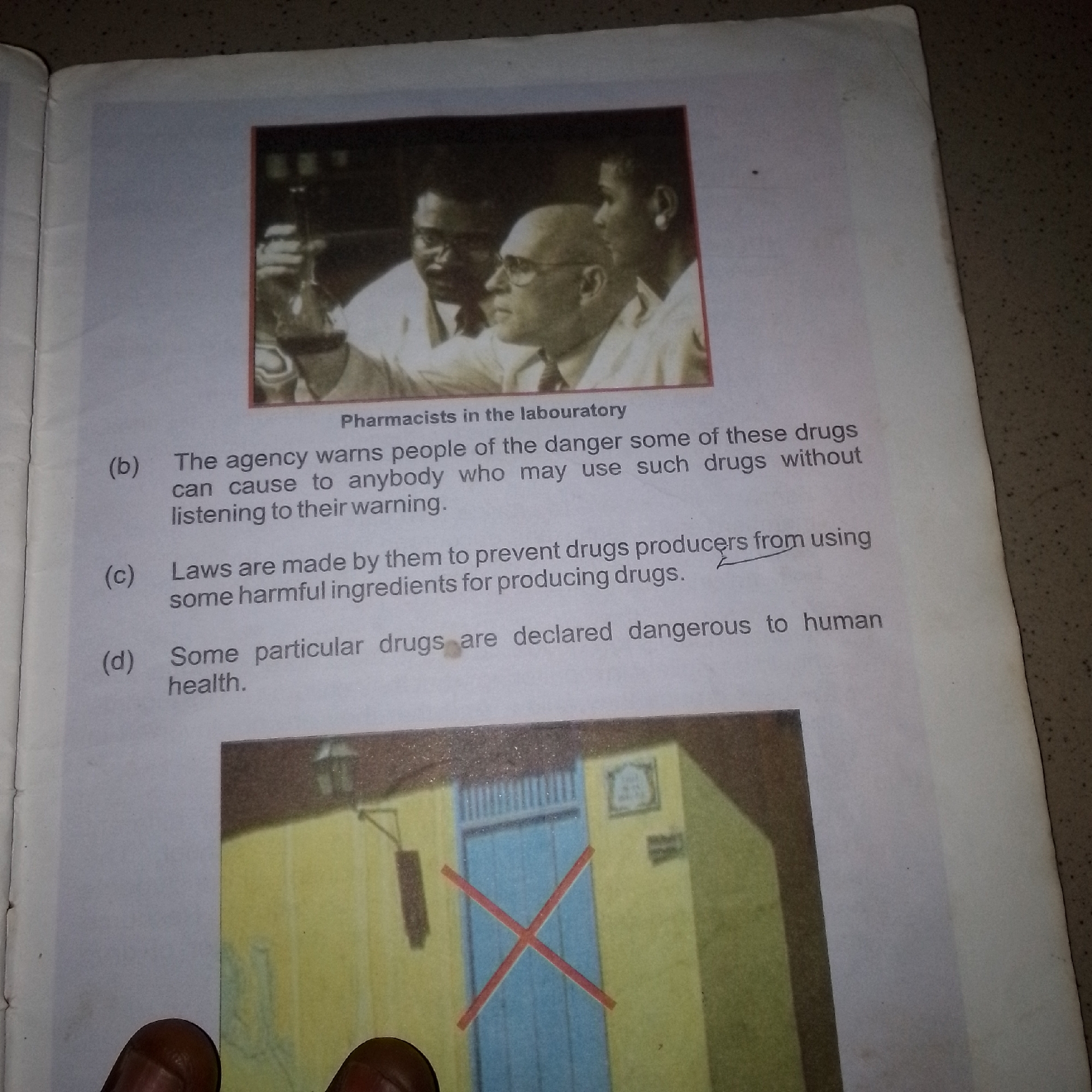 The image shows an open book with a photograph at the top and text below. The photograph is a black and white image of three pharmacists in a laboratory. They are wearing white lab coats and appear to be examining something. Below the photograph, there is text that reads "Pharmacists in the labouratory".
Following this is a list of numbered points, indicated by letters (b), (c), and (d). Point (b) states, "The agency warns people of the danger some of these drugs can cause to anybody who may use such drugs without listening to their warning." Point (c) reads, "Laws are made by them to prevent drugs producers from using some harmful ingredients for producing drugs." Point (d) states, "Some particular drugs are declared dangerous to human health."
At the bottom of the image, there is a color photograph of a building with a blue door that has a red 'X' marked on it. This photograph is cut off at the bottom. There are no visible landmarks or contextual elements in the image that would indicate a specific location in Jalingo, Nigeria.