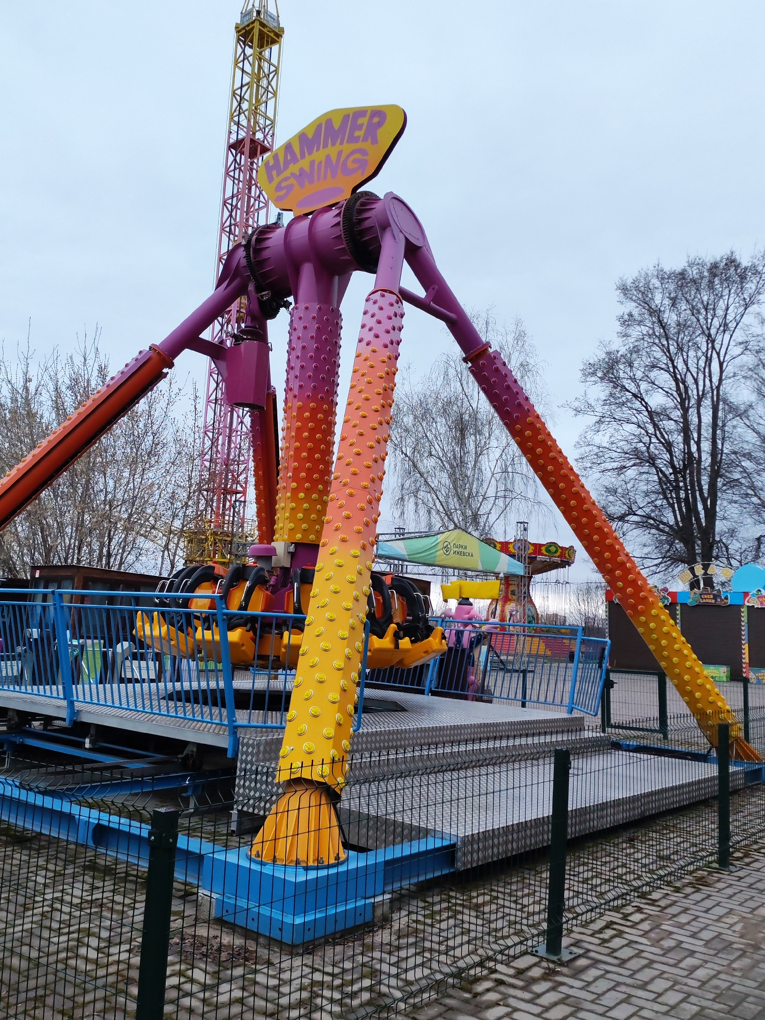 The image shows a "Hammer Swing" ride in a park in Izhevsk, Russia. The ride has a central purple structure with three arms extending outwards. These arms are decorated with a gradient of colors, from purple to orange to yellow, and studded with raised circles. The passenger seating area is attached to the ends of the arms and is primarily yellow and black. The ride is surrounded by a blue metal fence, with a grey metal platform on which the ride is based. In the background, other carnival rides and stalls can be seen. Bare trees and an overcast sky are visible above. A sign reading "Парки Ижевска" (Parks of Izhevsk) is partially visible in the background.