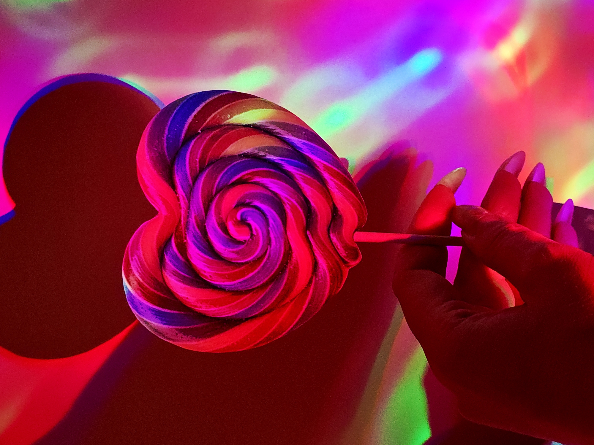 A vibrantly lit, close-up shot features a hand holding a heart-shaped lollipop. The lollipop has a spiral pattern of red, purple, and blue. The hand has long, pointed fingernails. The background is a mix of bright red, pink, purple, and green colors with blurred light patterns. There is a red shadow in the shape of a heart behind the lollipop.