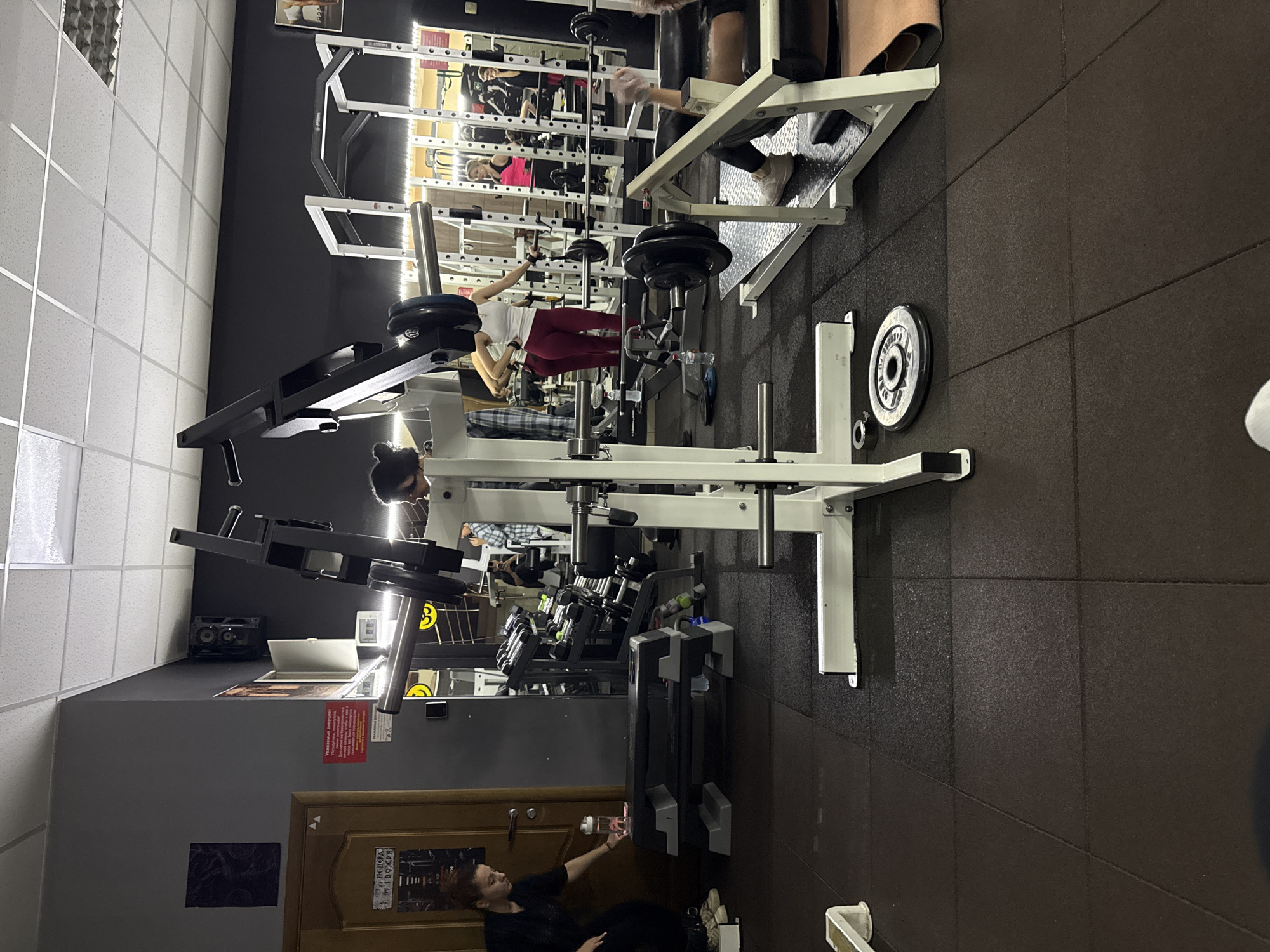 The image depicts the interior of a gym, specifically located in Nazran, Russia. The gym features a variety of weightlifting equipment, including a smith machine in the foreground and several squat racks and dumbbell racks in the background. The floor is covered with black rubber mats.

In the foreground, a woman with dark hair is visible on the left side of the image, wearing black clothing and sneakers. She is reaching for a clear water bottle with a pink liquid on a black step platform.

In the middle ground, several individuals are engaged in weightlifting activities. A woman in a white tank top and burgundy leggings is standing next to a squat rack, holding onto a barbell. To her right, another woman in a pink top and black leggings is also near a weight rack. Further in the background, other people are seen working out. One individual is lying on a bench press, with their arm raised.

The gym appears to be well-equipped, with weights, barbells, and machines strategically placed. The overall lighting in the gym is fluorescent. The walls are painted dark gray and black, with some posters and signage visible.