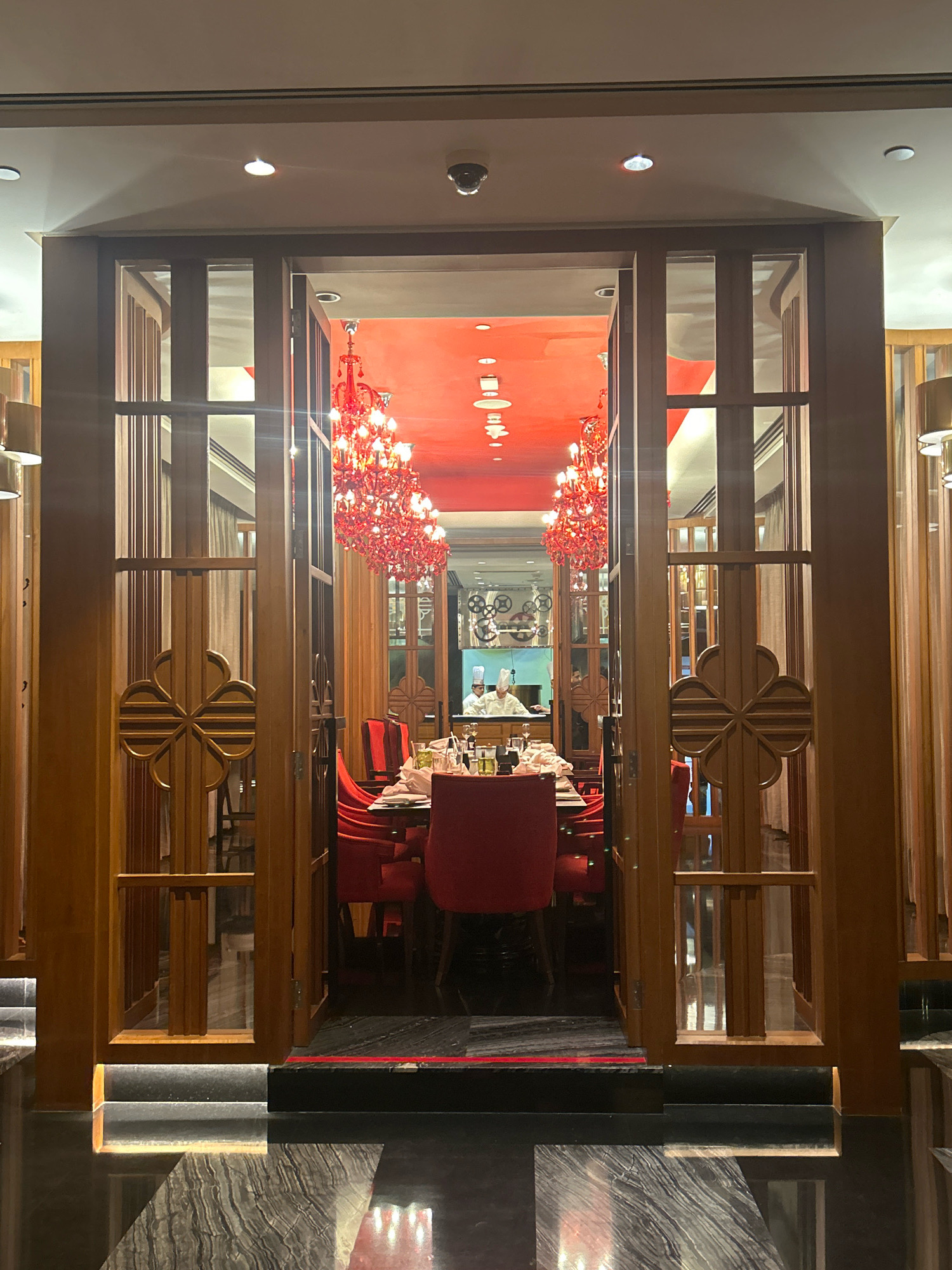 The image shows the entrance to a restaurant with a vibrant red ceiling and ornate red chandeliers. The entrance is framed by wooden doors and partitions with decorative carved panels. Inside, a long dining table is set with white tablecloths, plates, and glasses. Several red chairs are pulled up to the table. In the background, two chefs in white uniforms are visible in what appears to be a kitchen or prep area. The floor in the foreground is a polished dark stone with a reflective surface. The restaurant appears to be a high-end establishment. The location is identified as a restaurant in Hyderabad, India.
