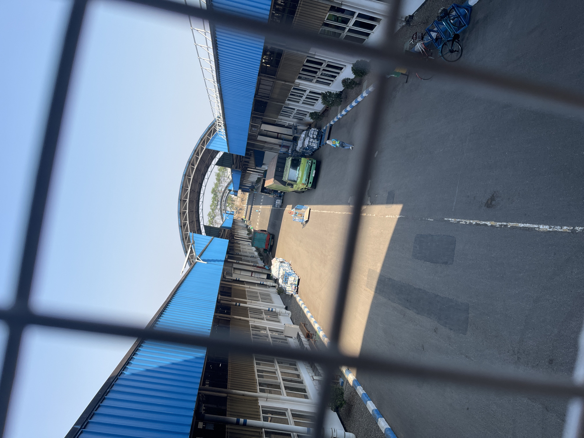 The image captures a sunlit outdoor scene behind a metal grate, likely at a logistics or industrial facility in Pelang, Indonesia. A long, paved roadway runs through the center of the frame, flanked by identical industrial buildings with vibrant blue, corrugated metal roofs and white, multi-paned windows. The buildings appear to be connected by an arched, open-air structure.
Several vehicles and objects are present on the roadway. A green truck is parked, and a pallet of what appears to be packaged goods is positioned beside it. A person wearing a light-colored shirt and dark pants, and a yellow safety vest, is walking away from the truck. Another person, similarly attired with a yellow vest and headscarf, is visible further down the road. A small, blue handcart or dolly is also on the pavement. Towards the right side of the frame, two bicycles are parked.
The ground is asphalt, with white lines marking lanes or parking areas. A blue and white striped border runs along the edge of the buildings. The sky is clear and blue, suggesting a bright day. Shadows are cast across the roadway from the buildings and structures. The overall impression is one of an active, operational industrial site.