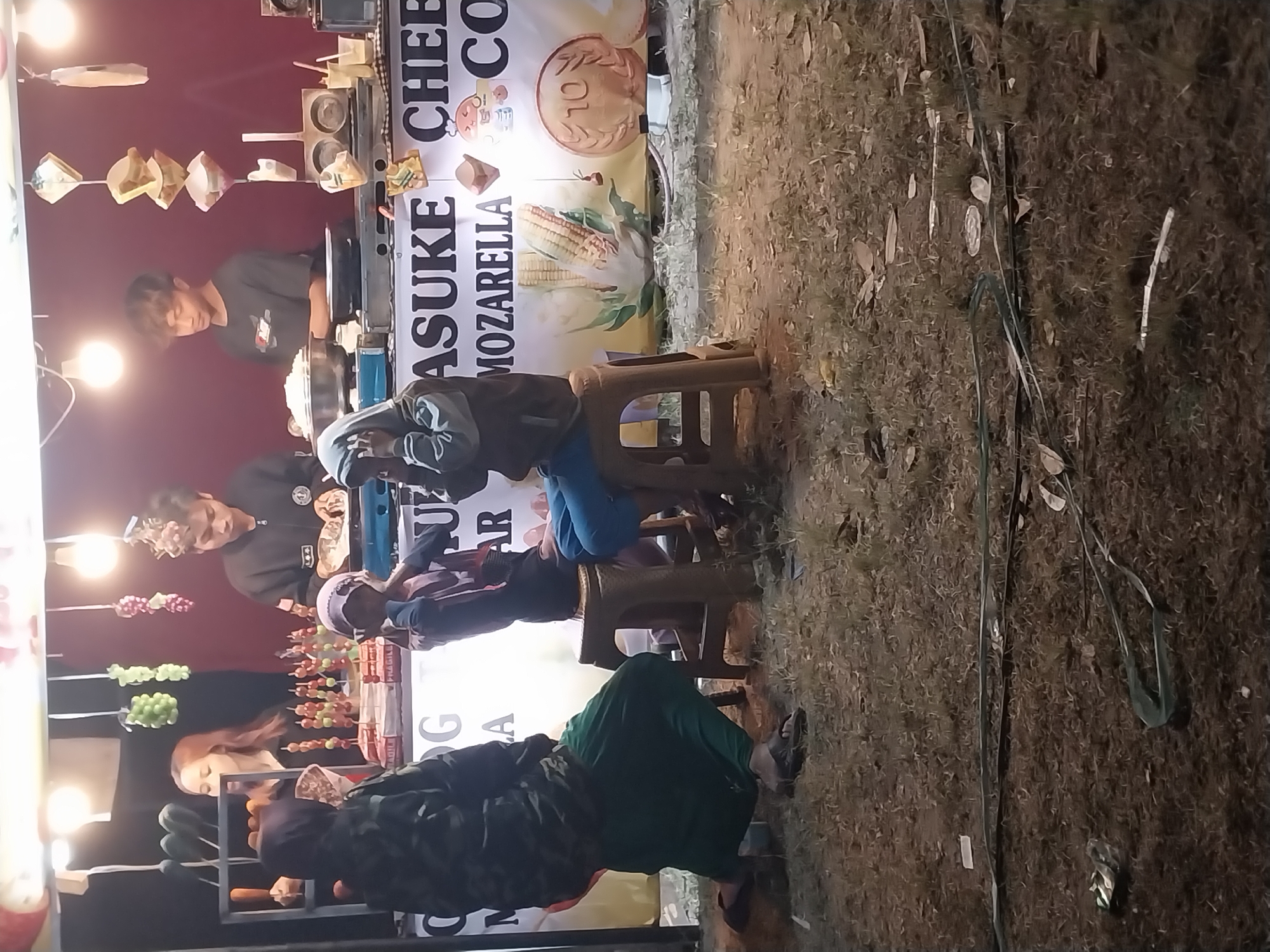 The image depicts an outdoor food stall at night, likely in Jiwut, Indonesia, based on the provided context. A female vendor with long, brown hair is visible behind a counter, holding what appears to be a food item on a stick. Two male vendors are also present, one wearing a dark jacket and the other a dark t-shirt with a graphic design. They are engaged in food preparation. Three customers are seated on stools in front of the stall. One customer wears a green sarong and a patterned jacket, another is obscured by a hijab, and a third is wearing a blue garment. A banner behind the vendors displays text including "ASUKE CHEESE MOZARELLA" and images of corn and a coin. The ground in front of the stall is covered with dry grass, fallen leaves, and some discarded items. Overhead lighting illuminates the scene.