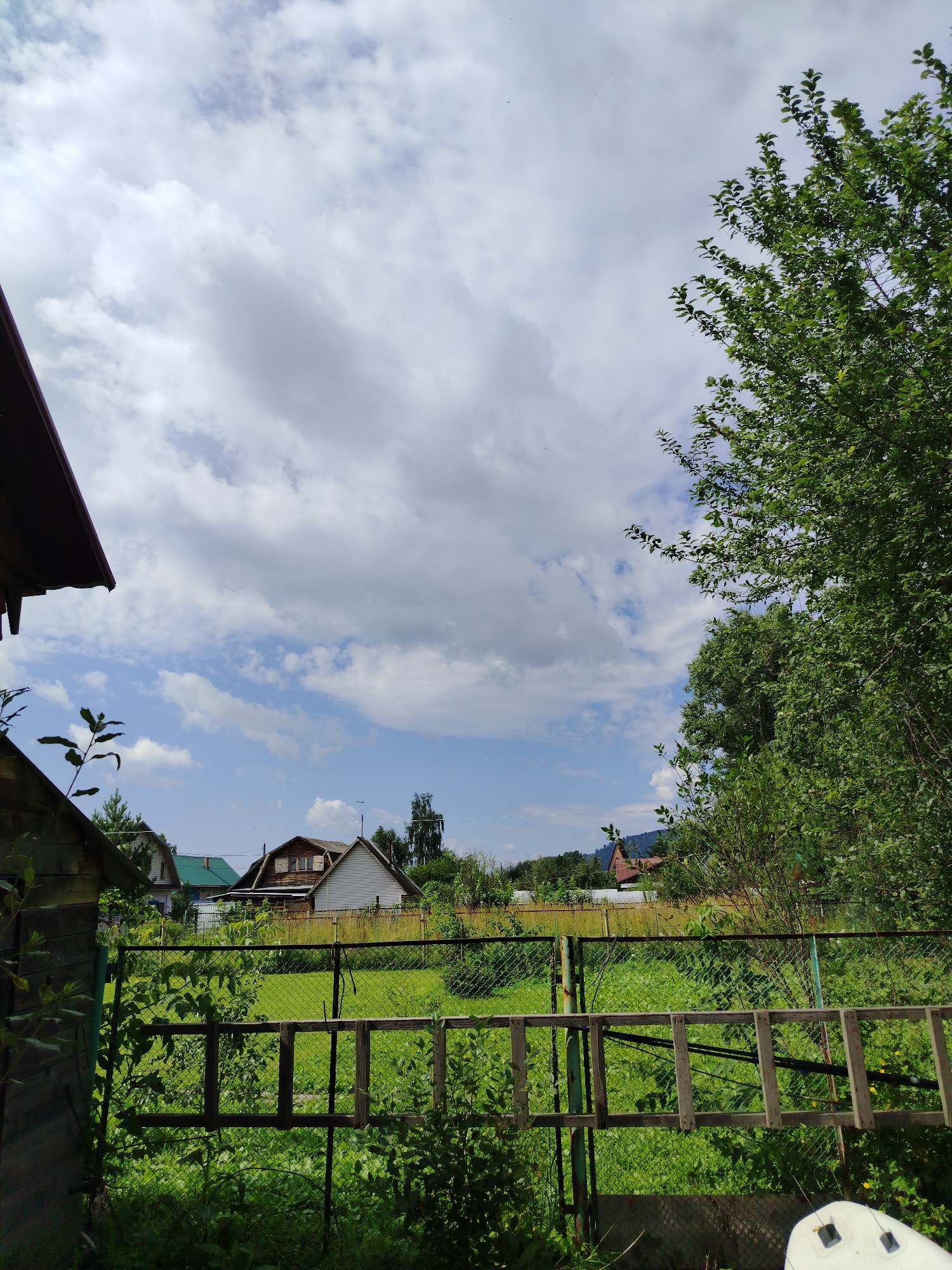 The image depicts a suburban or rural landscape with a focus on a fenced yard and several houses in the background. The foreground features a wooden structure on the left, possibly a shed or outbuilding, with a dark roofline visible. A chain-link fence runs across the middle ground, with a section of what appears to be a wooden ladder leaning against it. Beyond the fence, a green, overgrown area or field is visible. In the background, several houses with pitched roofs are scattered amongst trees. The sky is partly cloudy, with a mix of white and grey clouds against a blue sky. The overall impression is that of a residential area on a sunny day, possibly in the city of Kaltan, Russia, given the architectural style and vegetation. There are no people visible in the image.