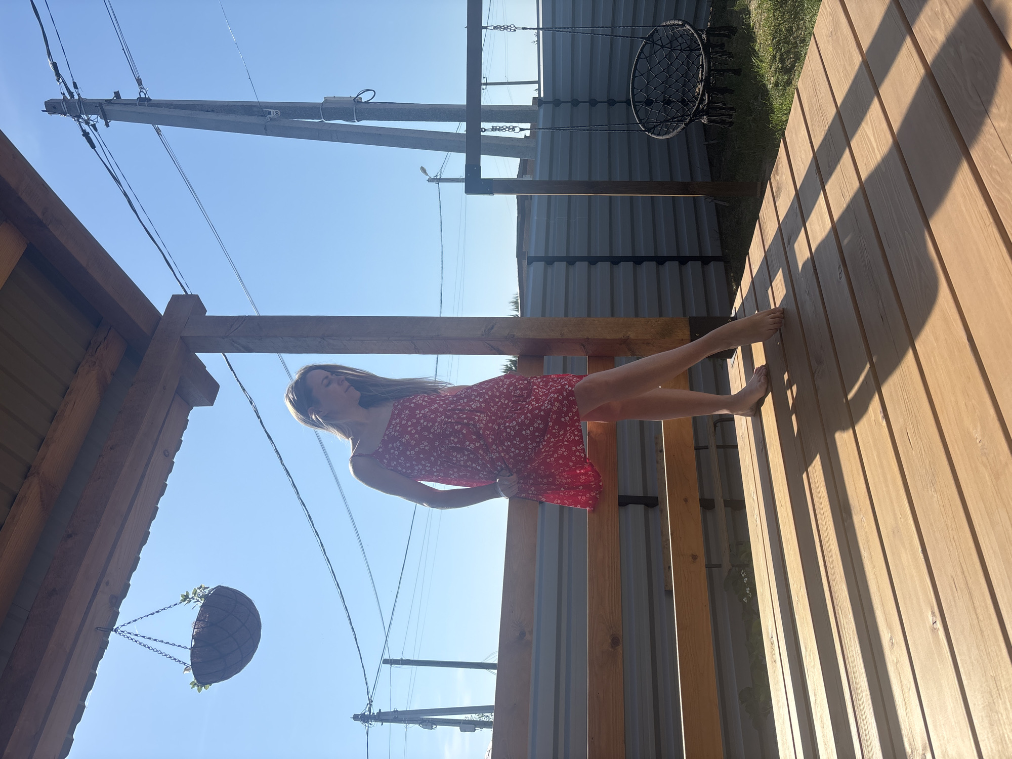 The image depicts a young woman in a short red dress, barefoot, standing on a wooden deck in what appears to be a backyard or outdoor space. In the background is a fence made of corrugated metal and several utility poles with power lines. A hanging pot and a circular swing are also visible. The photo seems to be taken outdoors under a clear, bright sky. This scene is located in the city of Chaikovsky, Russia.