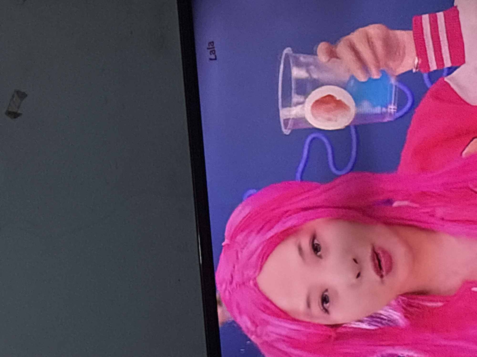 A close-up shot shows the face and upper body of a person wearing a bright pink wig and a jacket with pink and white stripes on the sleeve. The person has fair skin, dark eyes, and is looking slightly to their left. Their mouth is slightly open. To the right of the person, a hand is holding a clear plastic cup filled with a blue liquid. A white circular object is visible inside the cup. A purple cord snakes behind the cup. The background is a solid blue. The word "Lala" is visible in the upper right corner of the image. There is no identifiable information to place this image in Palimanan, Indonesia.
