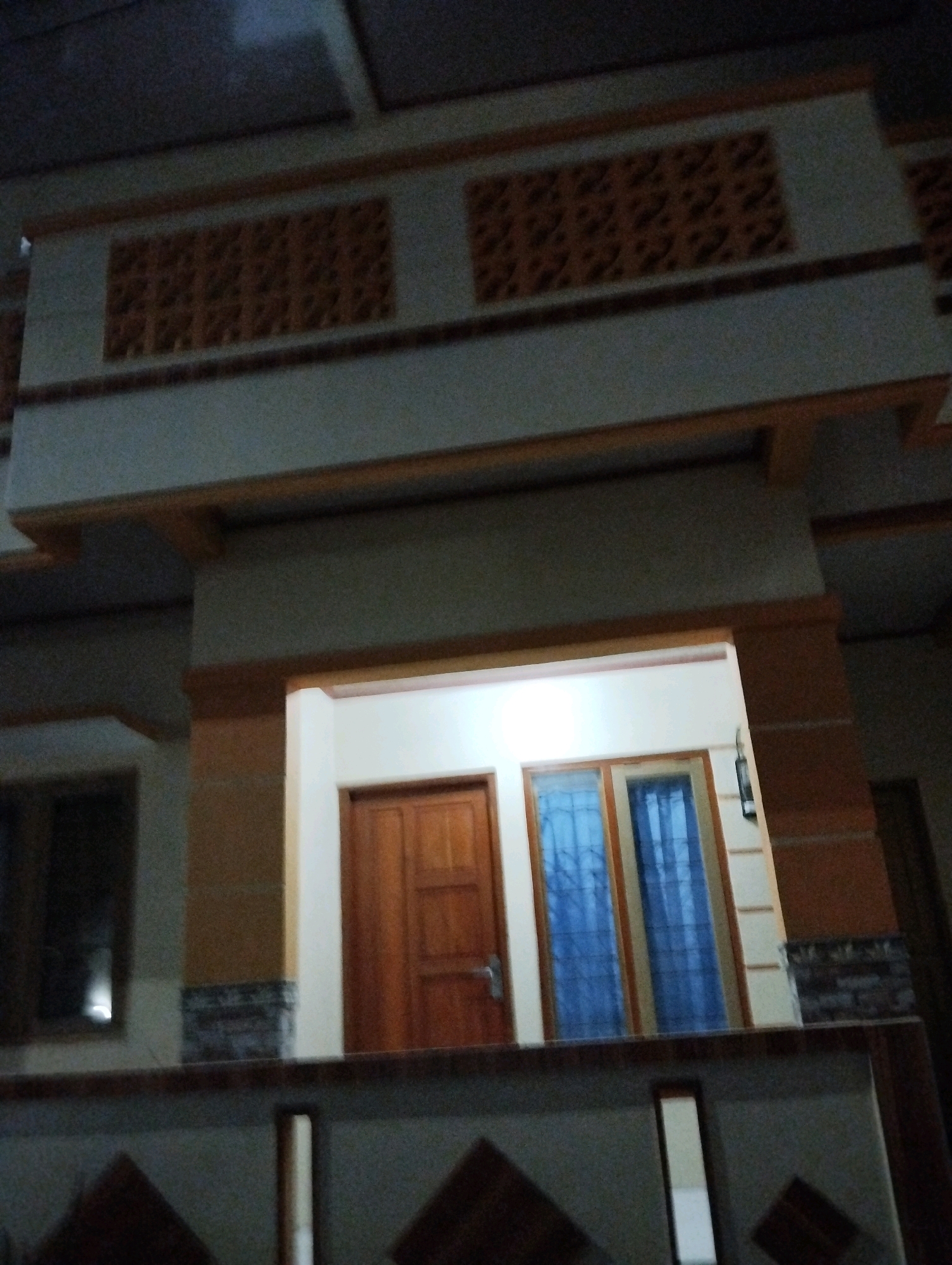 The image shows the exterior of a multi-story building, likely a house, in Salatiga, Indonesia. The building features decorative elements, including patterned vents on the upper level and a balcony or ledge in the foreground. The entrance consists of a wooden door flanked by two blue-tinted windows with decorative grilles. The lighting suggests it is either dusk or nighttime. The overall architectural style appears to be modern with some traditional influences. There are no people visible in the image.