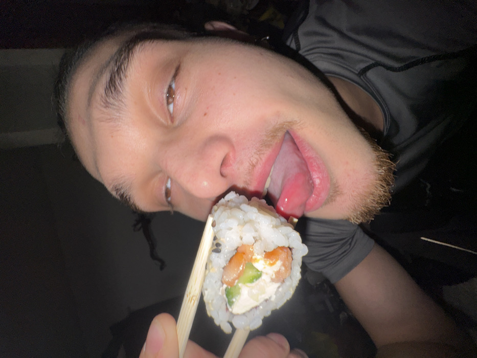 A close-up, dimly lit, low-angle indoor image depicts a male individual with dark hair pulled back, brown eyes, a faint nose piercing on the left nostril, and a short goatee. The individual's eyes are rolled slightly upward and to the right, and a bright pink tongue is extended towards a sushi roll. A hand, with visible thumb and index finger, uses light-colored wooden chopsticks to hold the sushi roll approximately 2-3 centimeters from the individual's mouth. The sushi roll is an inside-out type, covered in white rice and black sesame seeds, with a visible filling of a white creamy substance, orange-pink segments, and green strips. The individual is wearing a dark grey athletic-style shirt with visible seam stitching. The background is uniformly dark and indistinct, offering no discernible environmental features. There are no visual cues within the image to determine a specific location in the city of Sofia, Bulgaria.