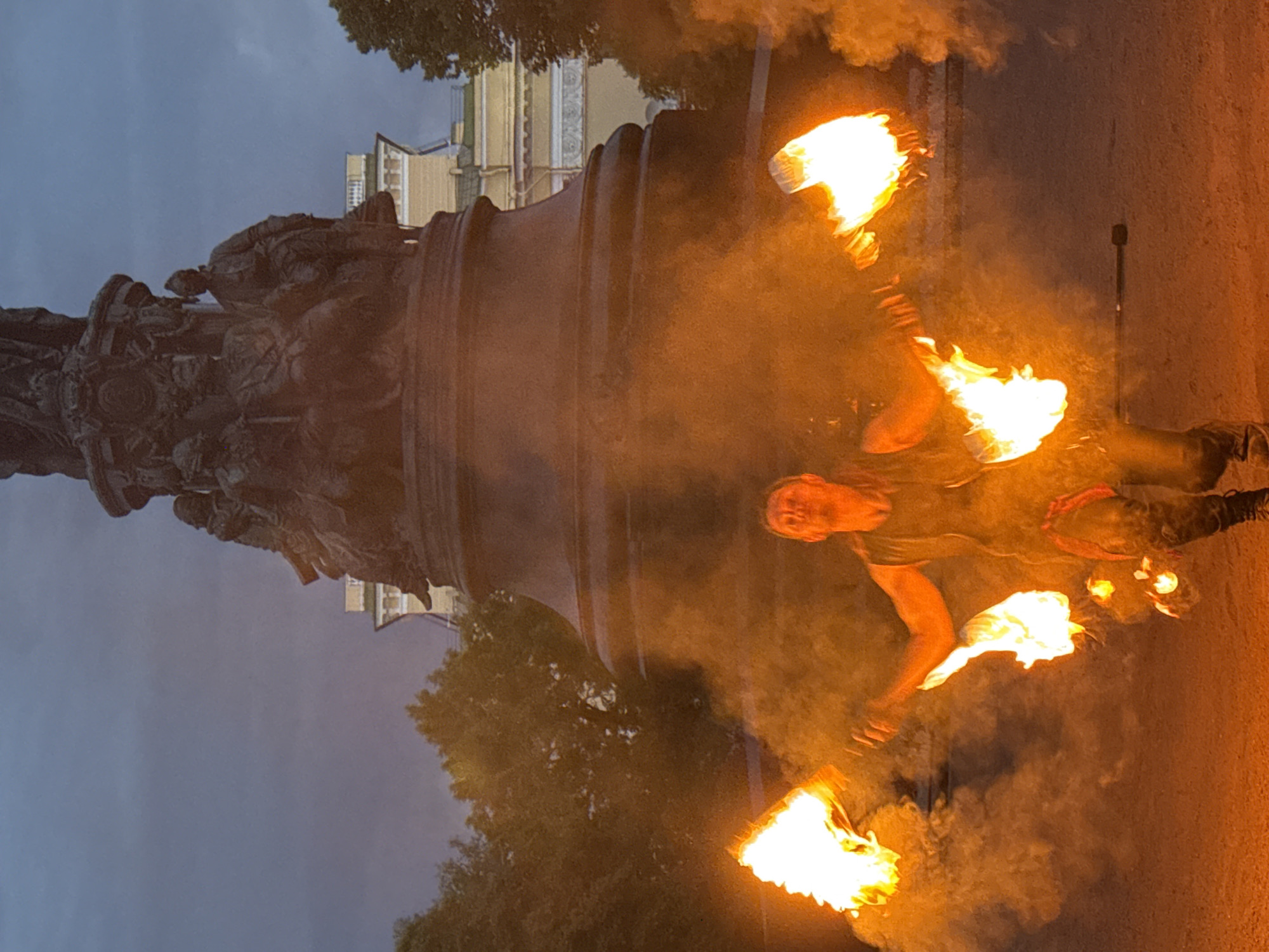 The image depicts a fire performance taking place in front of the Bronze Horseman monument in St. Petersburg, Russia. The central subject is a male performer wearing a sleeveless top and pants, actively engaged in manipulating two flaming torches. He is positioned in the foreground, surrounded by a cloud of smoke and the vibrant glow of the flames. Behind him, the imposing Bronze Horseman statue dominates the background, silhouetted against a dark, overcast sky. Trees and a light-colored building are also visible behind the monument. The ground in the foreground appears to be dark earth or asphalt. The overall atmosphere is dramatic, with the firelight casting strong shadows and illuminating the performer.