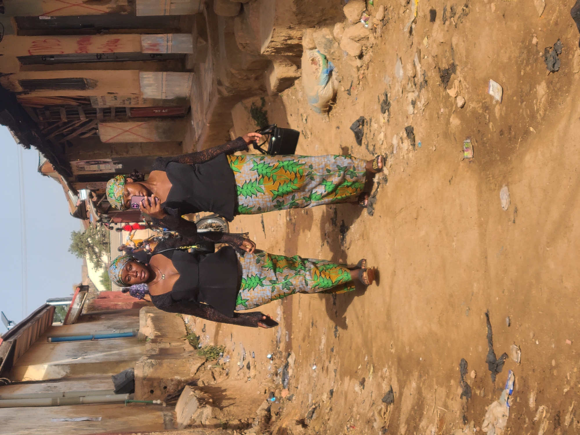 Two women stand side-by-side on a dirt road in Jos, Nigeria. Both are wearing similar attire: a black top with lace sleeves, a long, patterned skirt with green and yellow leaf-like designs, and head wraps. The woman on the left is holding a cell phone up to her face, seemingly taking a selfie or looking at something on the screen. The woman on the right is carrying a black purse. The road is cluttered with trash and debris. Buildings line both sides of the road, some appearing to be in disrepair. The sky is visible in the background.