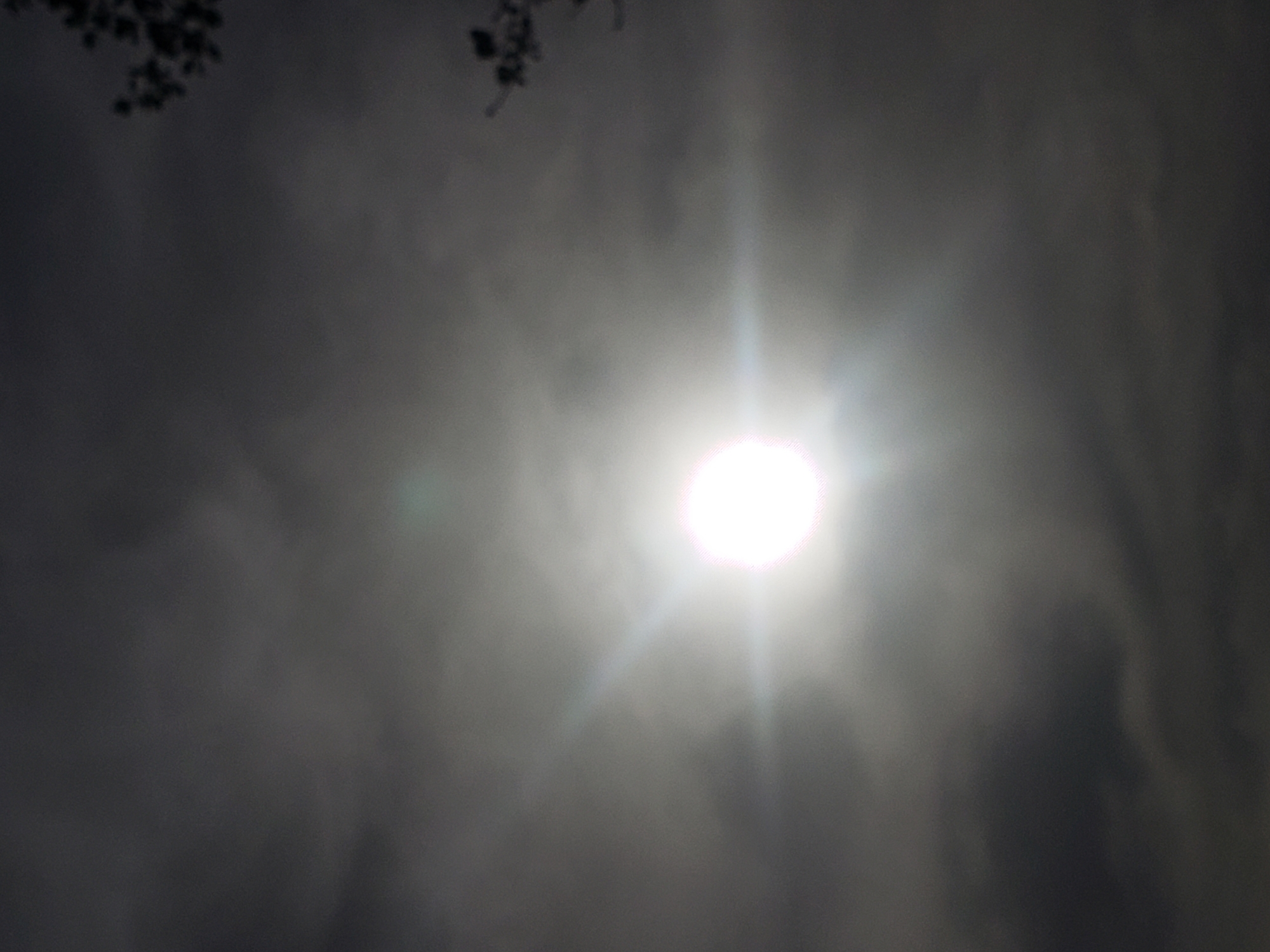 The image shows a bright light source, likely the sun, obscured by a layer of clouds or mist. The sky is predominantly gray and dark, suggesting overcast conditions. A few dark, indistinct shapes, possibly tree branches or foliage, are visible in the upper left corner. The location is Adankolo, Nigeria.