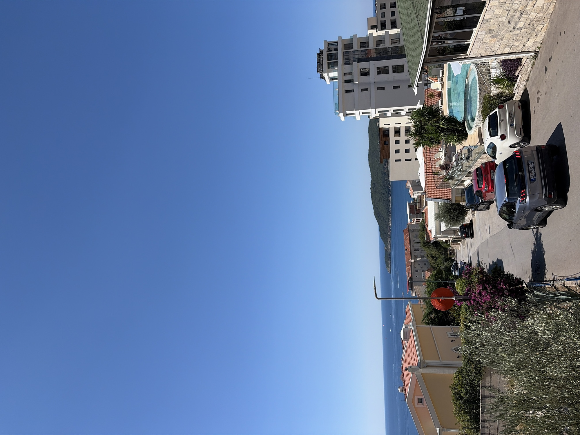 This is a wide-angle, high-angle shot of a coastal town on a clear, sunny day, likely in Bečići, Montenegro. The sky is a vibrant, cloudless blue, filling the upper half of the frame. Below the sky, the deep blue Adriatic Sea stretches to the horizon, dotted with distant boats and a verdant, hilly coastline.
The foreground is dominated by a narrow, paved street winding downhill. Several cars are parked or driving on the street. Closest to the viewer is a silver Peugeot SUV, followed by a white Alfa Romeo, a red hatchback, and a black car further up the street.
To the left of the street, a yellow house with a terracotta roof stands, partially obscured by lush green foliage. To the right, a modern white building with multiple stories, glass balconies, and rooftop solar panels is prominent. Adjacent to this building is a swimming pool area with lounge chairs and umbrellas, and a circular hot tub. A building with a green roof and stone facade is situated to the far right.
Further up the street, more buildings with red-tiled roofs and Mediterranean architectural styles are visible. A prominent street lamp with a modern design stands near a cluster of vibrant purple bougainvillea flowers. The overall impression is a picturesque coastal resort town bathed in bright sunlight.