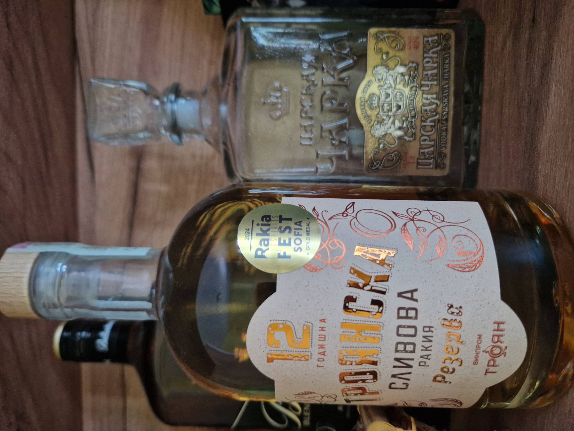 The image displays three bottles of alcoholic beverages arranged on a wooden surface against a wooden background.
In the foreground, slightly left of center, is a clear glass bottle containing a pale amber liquid, characteristic of a spirit. The bottle is sealed with a light-colored wooden stopper. Its main label is irregular in shape, off-white, and features prominent gold-colored Cyrillic text that reads "12 ГОДИШНА" (12-year-old), "ТРОЯНСКА СЛИВОВА РАКИЯ Резерва" (Troyan Plum Rakia Reserve), and "ВИНПРОМ ТРОЯН" (Vinprom Troyan) at the bottom. Copper-colored foil outlines of fruit, possibly plums, and decorative swirling patterns adorn the label. A circular gold medal sticker is affixed to the upper right of the label, bearing the text "2024 Rakia & Spirits FEST SOFIA GOLD MEDAL".
To the right and slightly behind the prominent bottle stands an empty, clear glass bottle or decanter, rectangular in shape, topped with a square-cut, ornate glass stopper. The body of this bottle is embossed with Cyrillic text reading "ЦАРСКАЯ ЧАРКА" (Tsarskaya Charka) beneath a crown symbol. A rectangular gold-colored label with black borders is affixed to its front, depicting a crest with two rampant lions supporting a shield, crowned at the top. Text on this label includes "ЗОЛОТАЯ GOLD" (Golden Gold), "ВОДКА ЦАРСКАЯ ЧАРКА" (Vodka Tsarskaya Charka), and "VODKA TSARSKAYA CHARKA", with "40%" visible on the upper right.
In the blurred background on the left, a dark-colored glass bottle is partially visible. It has a black cap with a gold band and a portion of a white label with dark text, possibly "BLEN" or "BLEND".
All bottles rest on a horizontally grained wooden surface, appearing to be a shelf or tabletop, with a vertically grained wooden panel forming the background. Both wooden surfaces are of a medium brown hue with visible natural grain patterns. The lighting suggests an indoor setting, with illumination primarily from the front, highlighting the labels and the contents of the foreground bottle. No individuals are present.