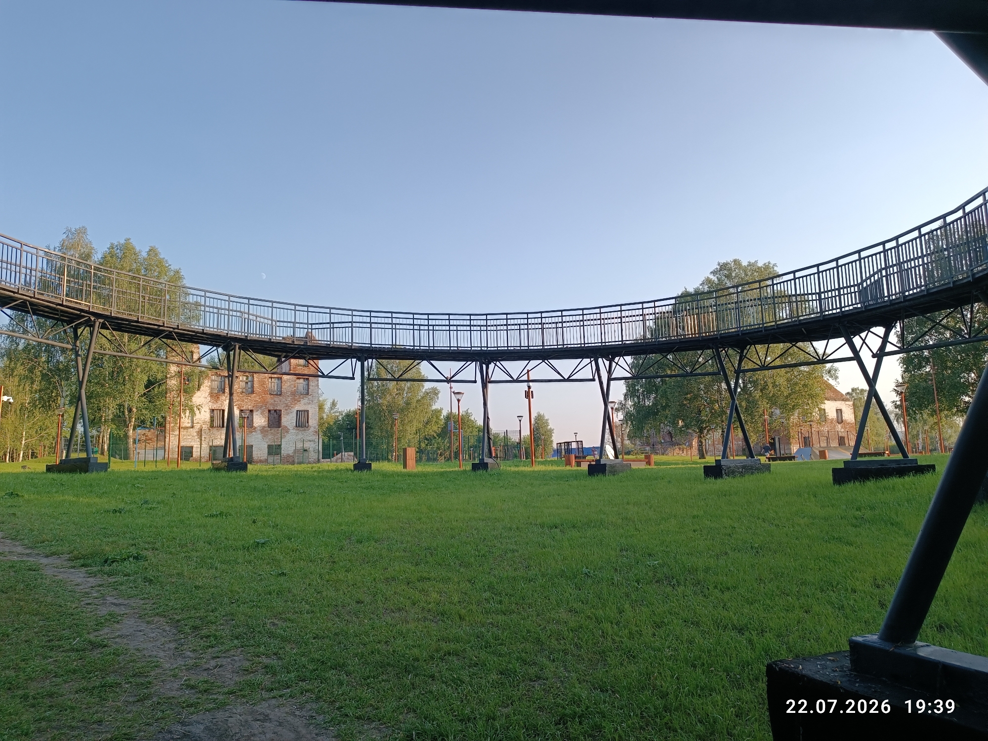 The image depicts a curved metal walkway elevated above a grassy field, with an old brick building visible in the background. The walkway has a metal railing and is supported by metal structures. Several lampposts are scattered across the field. Trees surround the building and the walkway. The sky is clear and blue. The lower right corner of the image shows a timestamp indicating July 22, 2026, 19:39. The location is identified as Verkhoturye, Russia. There are no people visible in the image. The scene appears to be a park or outdoor recreational area.