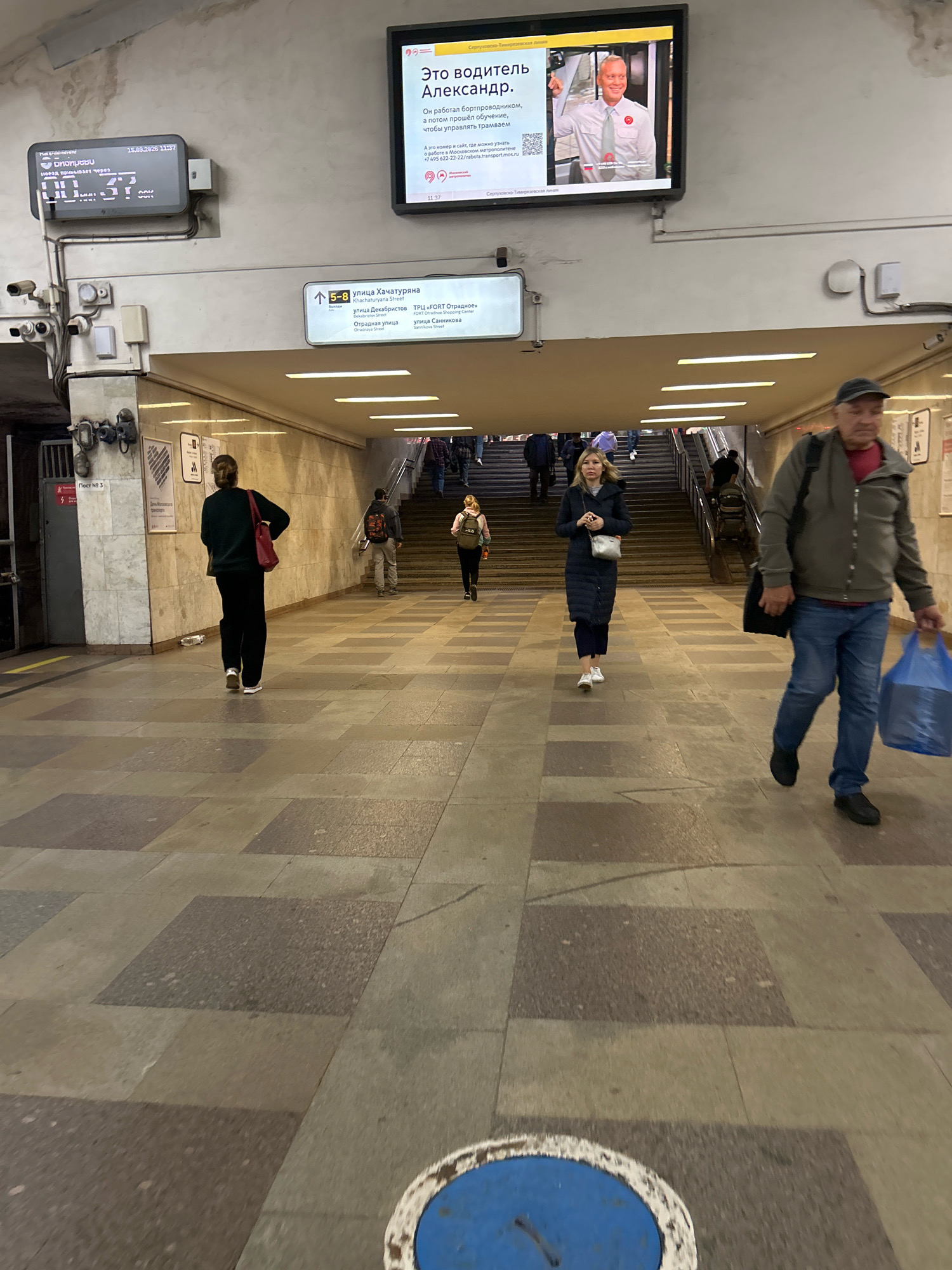 The image captures a busy underground passage in Moscow, Russia, likely a metro station, as indicated by the signage and electronic displays. In the upper left, a digital display shows "15.02.2026 11:37" alongside a timer. To its right, a large screen displays an advertisement featuring a man in a uniform and text in Russian, promoting employment as a driver.
Several people are seen navigating the passage. In the foreground right, a man in jeans and a grey jacket walks towards the viewer, carrying a blue plastic bag. Further down the passage, a woman in a dark coat walks away from the camera. Others are visible in the mid-ground and background, some ascending a set of stairs leading upwards, suggesting an exit or another level of the station. The environment is characterized by tiled floors and walls, with overhead lighting illuminating the space. There are also various signs and service elements visible, such as security cameras and electrical boxes. The overall scene depicts a typical day in a public transportation hub.