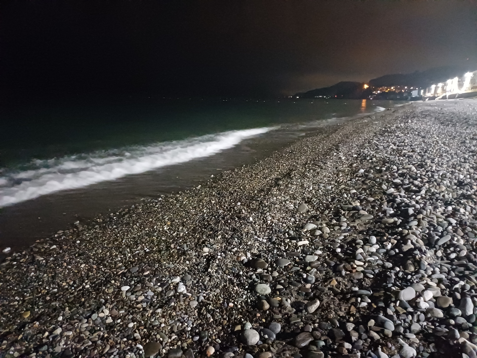 This nighttime image depicts a pebble beach scene in Sochi, Russia, devoid of people. The foreground is dominated by a wide expanse of dark, wet and dry pebbles in various shades of grey, white, and brown. Moving into the midground, the dark sea meets the shore, with multiple elongated white foam lines indicating breaking waves. The water itself appears a deep greenish-blue, reflecting minimal ambient light. The background reveals a distant coastline silhouetted by dark mountains, with numerous scattered artificial lights outlining the shoreline and a city or town. A diffused orange-yellow glow in the sky above the distant land indicates light pollution from the urban area, with a concentrated cluster of brighter white lights visible on the far right, suggesting illuminated structures or a promenade.