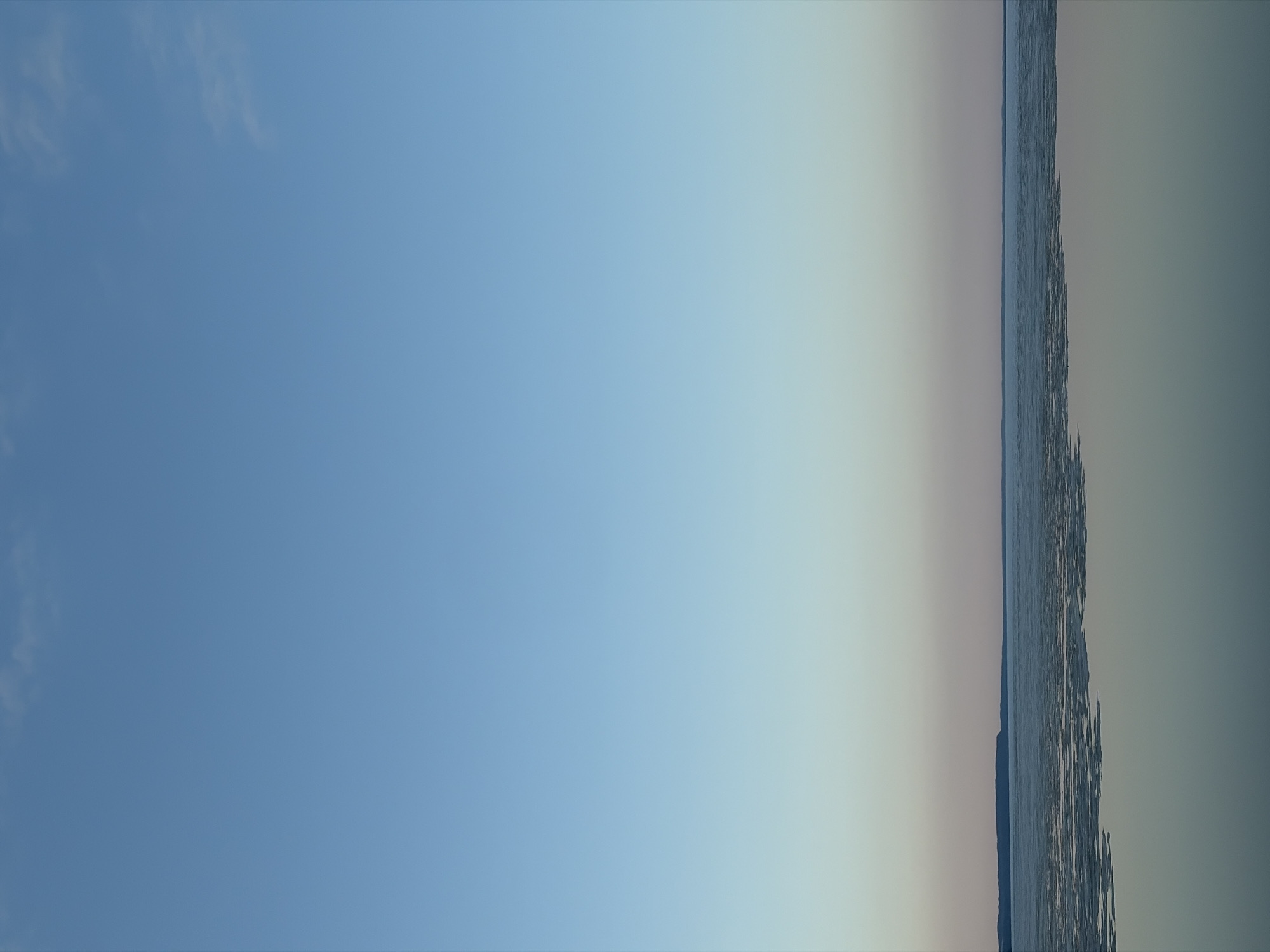 The image presents a horizontal view, with the right portion depicting a body of water. The water appears textured by wind or small waves. Adjacent to the water, the sky transitions from light to dark. The left side predominantly features a clear blue sky, subtly tinted with lighter shades toward the center. The location is Chaikovsky, Russia, based on the image's origin. There are no people or discernable objects visible other than the water and sky.