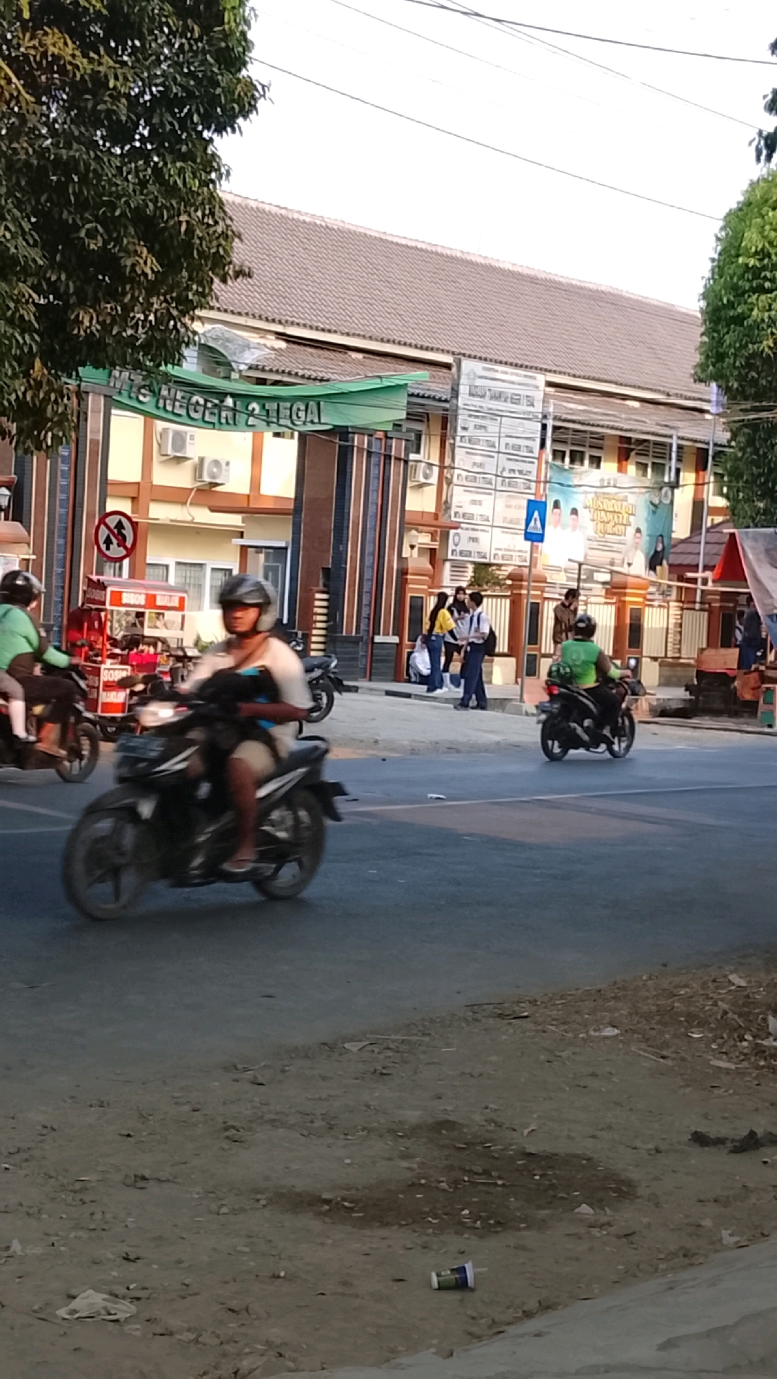 The image depicts a street scene in Slawi, Indonesia, likely in front of an educational institution, identified by a green banner overhead reading "MTS NEGERI 2 TEGAL." In the foreground, a person is riding a black motorcycle, captured in motion blur. To the left, another motorcycle is also in motion blur, with a rider wearing a green shirt and a helmet, and a red food cart with the word "SOSIS" visible. In the background, several individuals are standing on a sidewalk near the building. The building itself has a tiled roof and cream-colored walls with brown and blue decorative columns. A street sign with a red circle and a diagonal line is visible on the left. The ground surface is a mixture of asphalt and dirt, with some litter, including a disposable cup, scattered near the edge. The sky is overcast, suggesting it might be late afternoon or early morning.