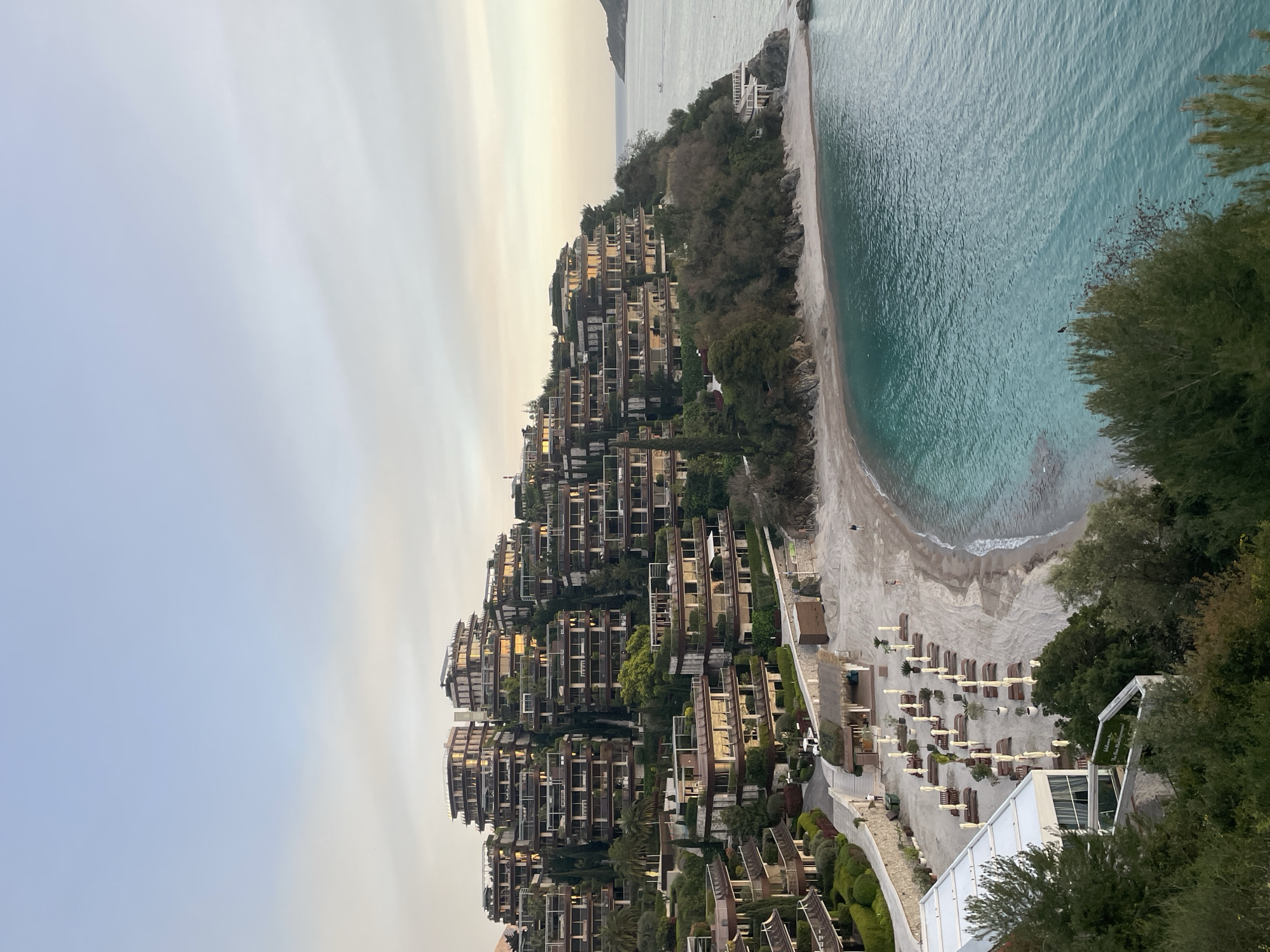 An aerial view of a multi-tiered hotel complex on a hillside in Budva, Montenegro, adjacent to a sandy beach. The resort features numerous balconies and terraces. Below, the beach contains rows of lounge chairs and umbrellas. A turquoise sea borders the beach. The sky is overcast.