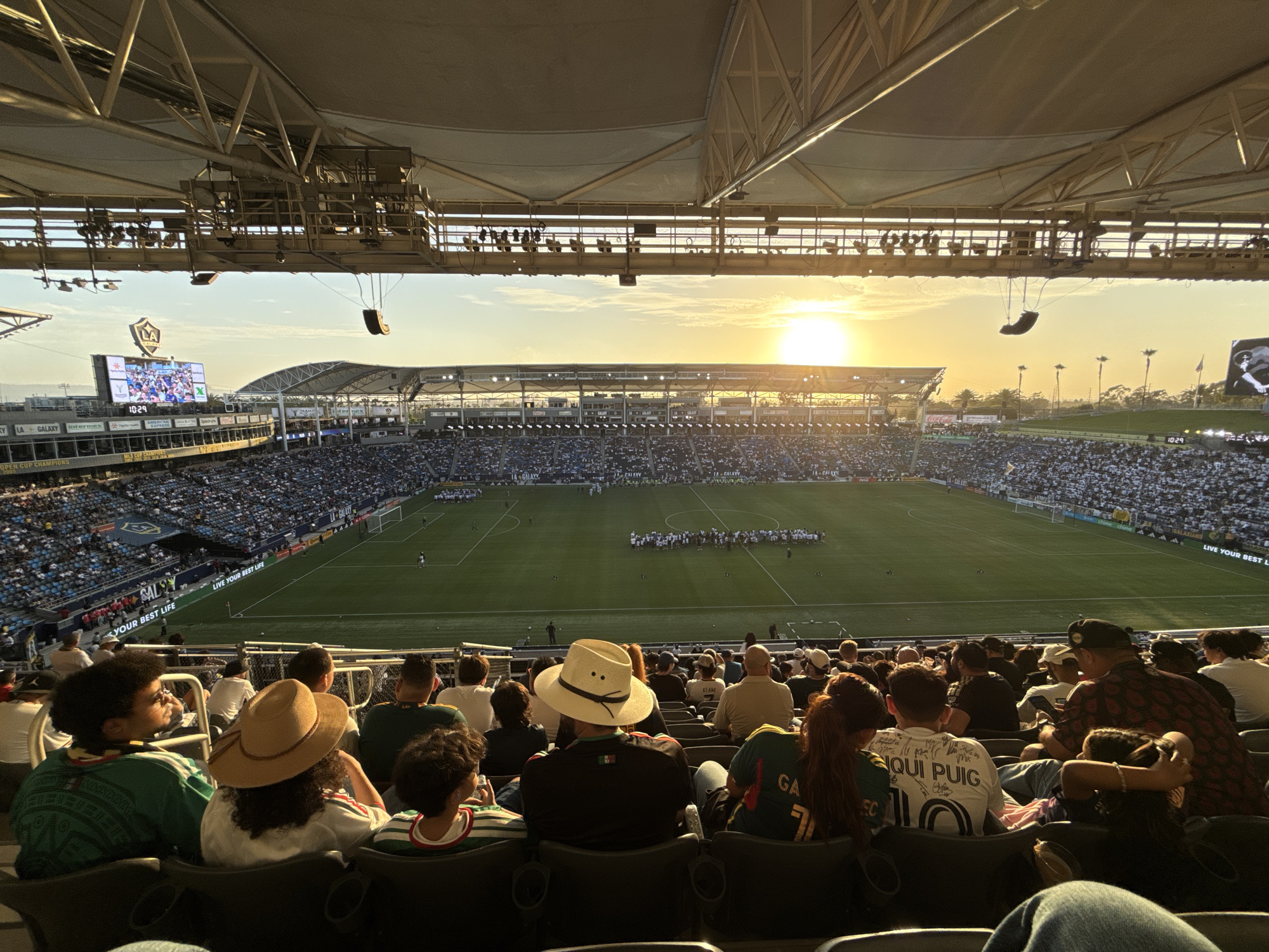 In the city of Carson, United States of America, a soccer match is underway at Dignity Health Sports Park. The image captures a wide view of the stadium, focusing on the stands filled with spectators. The setting sun casts a warm glow over the scene, illuminating the field and the stadium's architecture. In the foreground, a group of fans are seated, their backs to the camera, as they watch the game. Their attire and body language suggest engagement and enjoyment of the event. The stadium itself appears modern and well-maintained, with a large screen displaying game information. The overall atmosphere is one of a typical sports event, with a blend of athletic competition and community gathering.