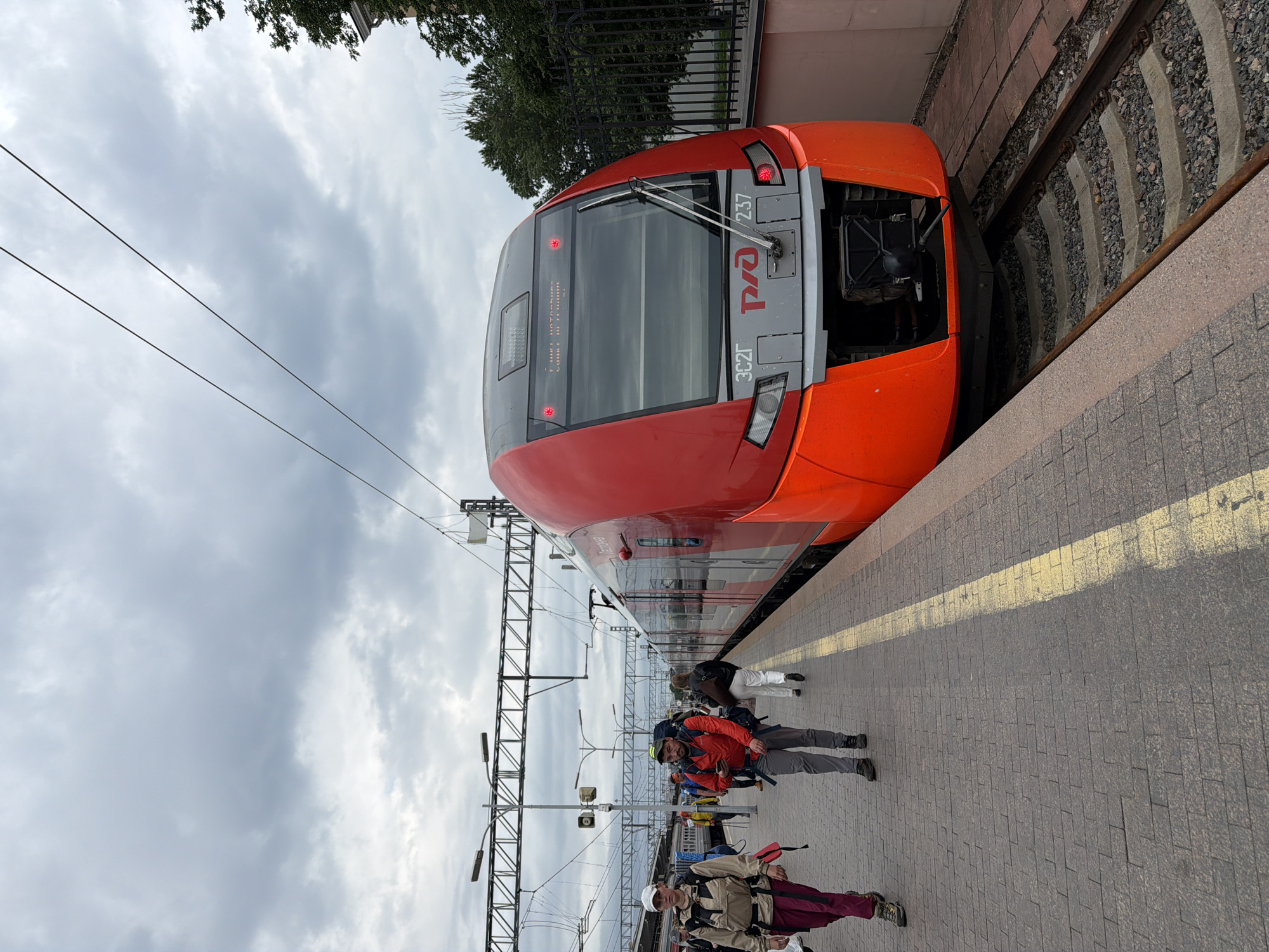 This image captures a red and gray train, identified by the "РЖД" logo and the number "237" on its front, stopped at a train station platform in Saint Petersburg, Russia. The sky is overcast with clouds. Several people are visible on the platform. In the foreground, a woman wearing a light-colored jacket and dark pants with a backpack walks to the left. To her right, a man in a red jacket, gray pants, and a backpack is walking towards the viewer. Behind them, other passengers are also seen on the platform. The train is a modern electric multiple unit, indicated by its sleek design and the overhead electrical lines. The platform is paved with bricks, and a yellow line marks the edge of the platform. A black metal fence is visible to the right of the train, bordering a grassy area with trees. The overall scene depicts a typical day at a busy train station in a Russian city.