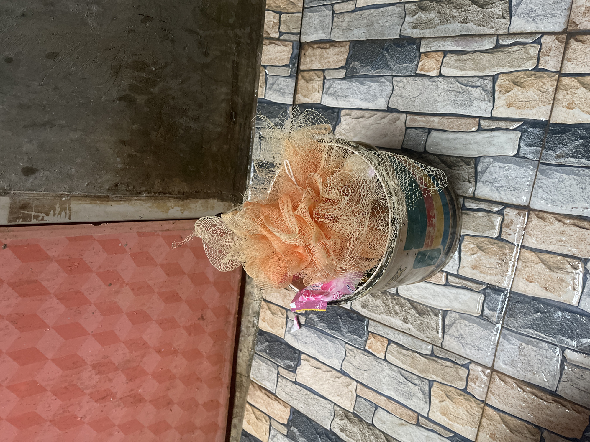 An orange pouf sits in a tin bucket on a tiled floor. The tin bucket is a repurposed food or paint container with colorful bands of red, yellow, green, and blue around its circumference. The pouf itself is made of netting and appears to be a bath accessory, with a small pink packet, possibly soap or lotion, attached to its side. The floor is covered in rectangular tiles arranged in a brick pattern, with a mix of beige, brown, grey, and black stones. To the left of the pouf is a red door with a diamond-shaped pattern. To the right of the door is a textured grey wall, possibly concrete, with some water stains and markings. The scene appears to be in an outdoor or semi-outdoor area, likely a bathroom or entryway. The lighting suggests it is daytime. This scene is located in Kebumen, Indonesia.